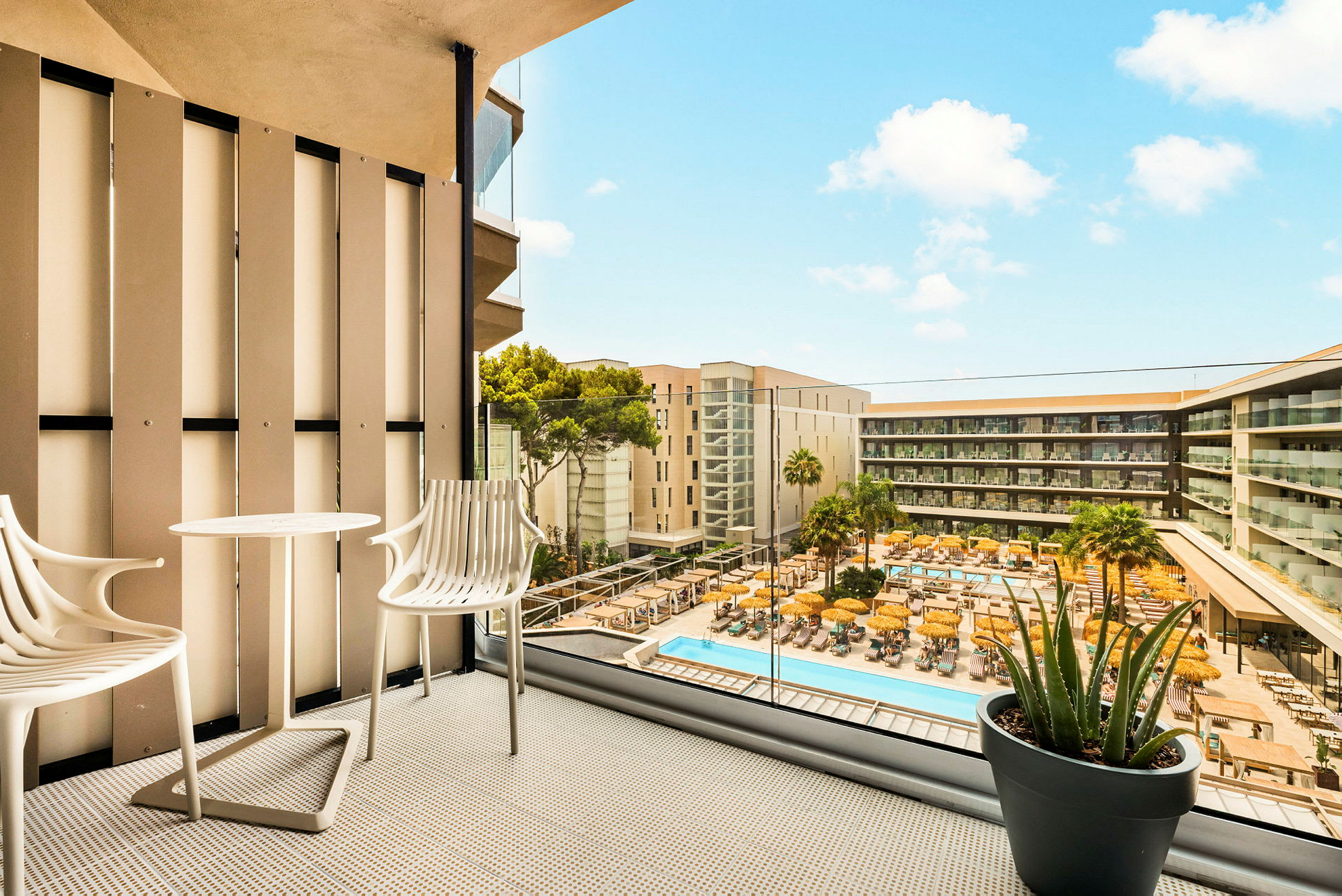 Utsikt från en balkong med två stolar och ett bord, med vy över en pool och hotellbyggnader. (AI-genererad text)