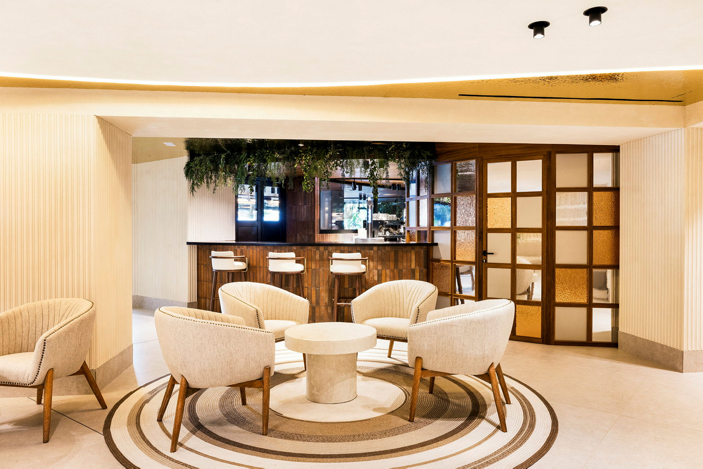Ett modernt hotellområde med en sittgrupp av mjuka fåtöljer runt ett runt bord och en bardisk i bakgrunden. Inredningen är ljus och elegant med växtdekorationer. (AI-genererad text)