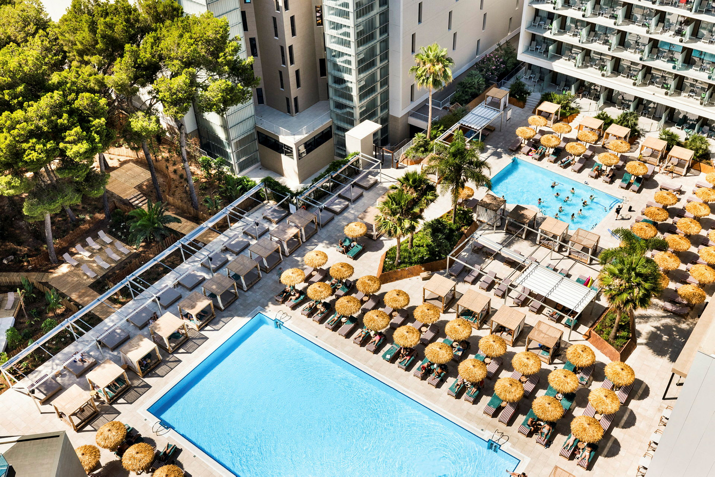En hotellpool med solstolar och parasoller omgiven av byggnader och träd. (AI-genererad text)