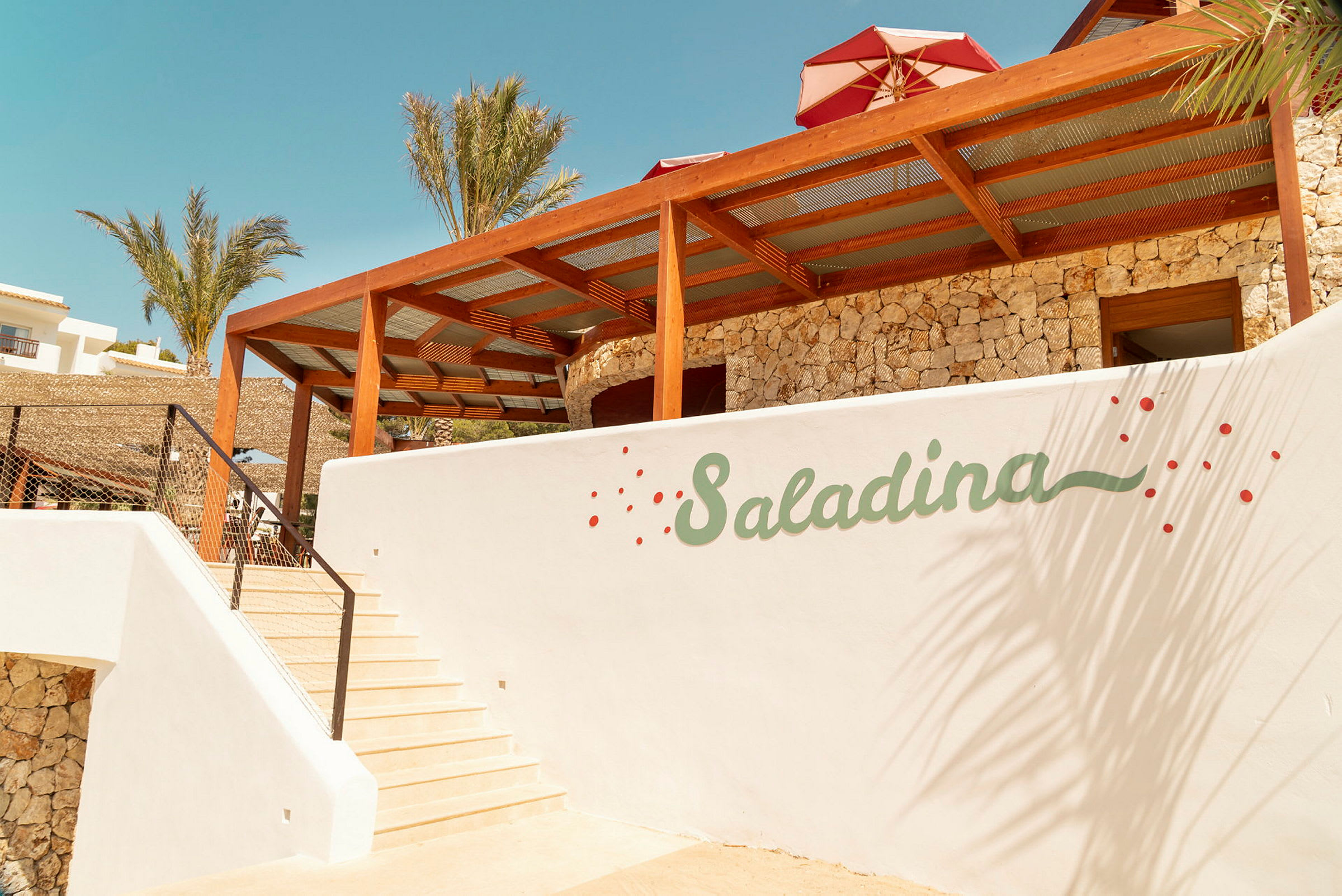 Vit byggnad med trappor, logotypen "Saladina" på väggen och palmträd i bakgrunden. (AI-genererad text)