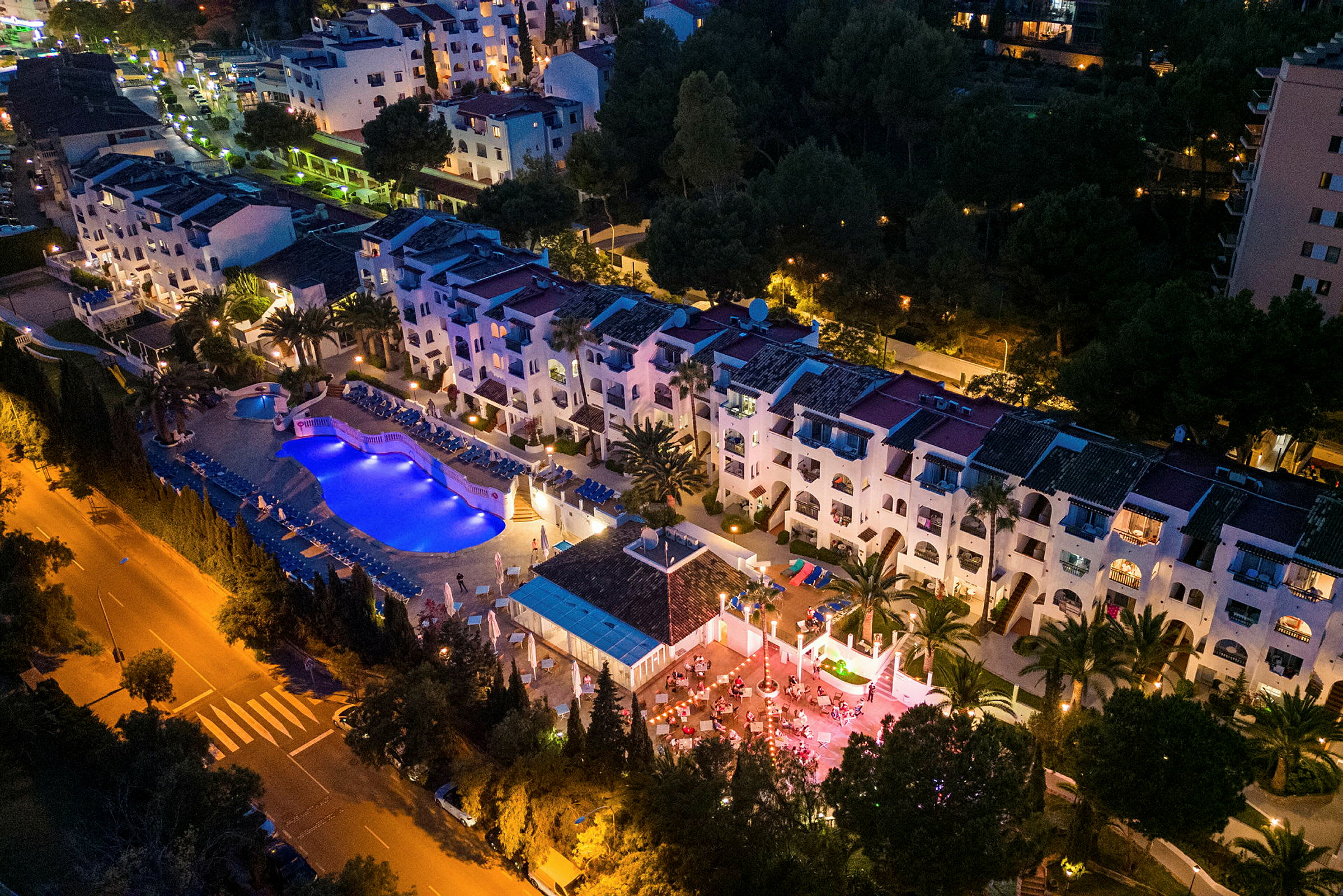 Flygbild av en upplyst hotellanläggning med pool och terrass under kvällstid. (AI-genererad text)