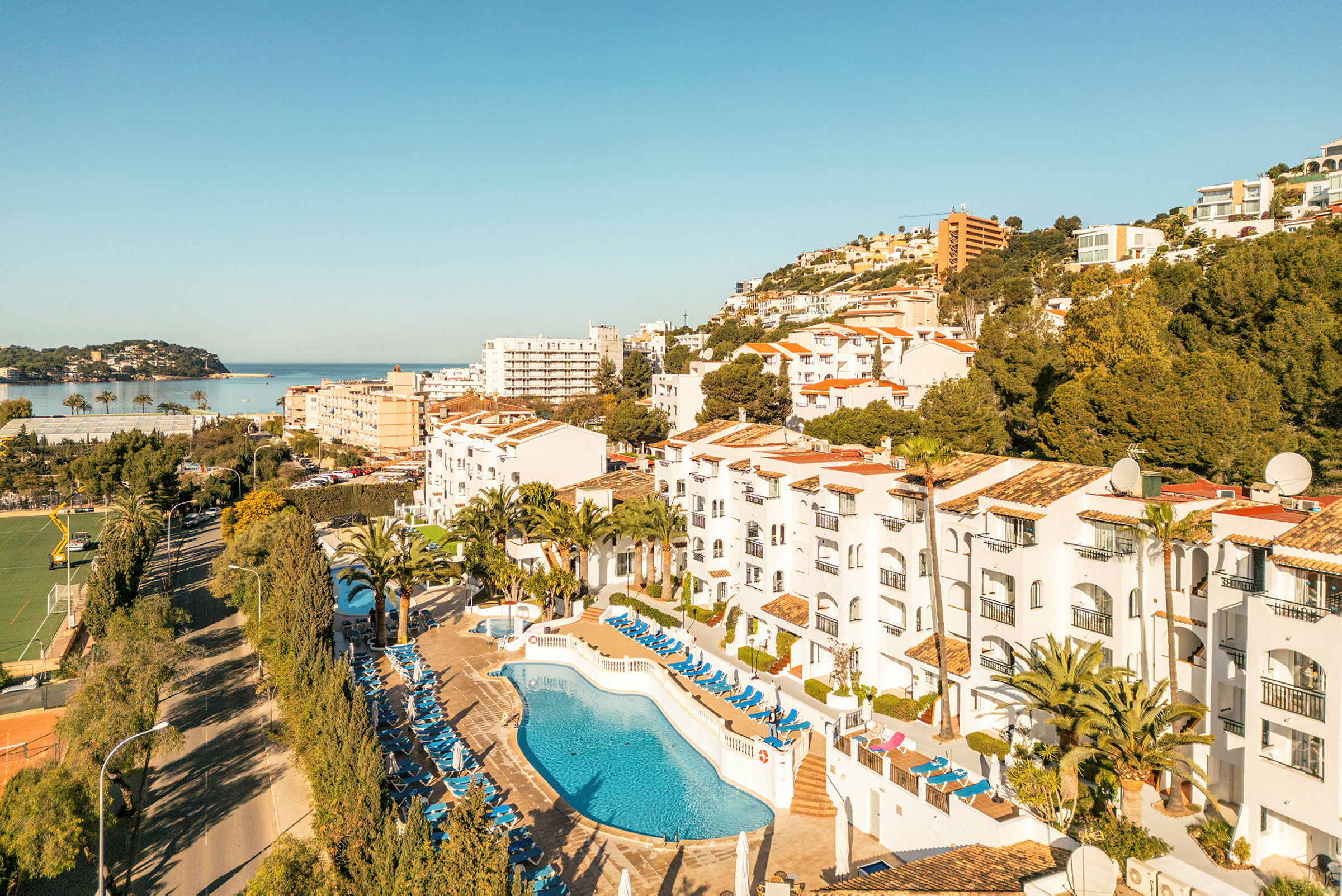 Bild av hotellområde med pool och solstolar, omgivningar med träd och byggnader samt en vy mot havet. (AI-genererad text)