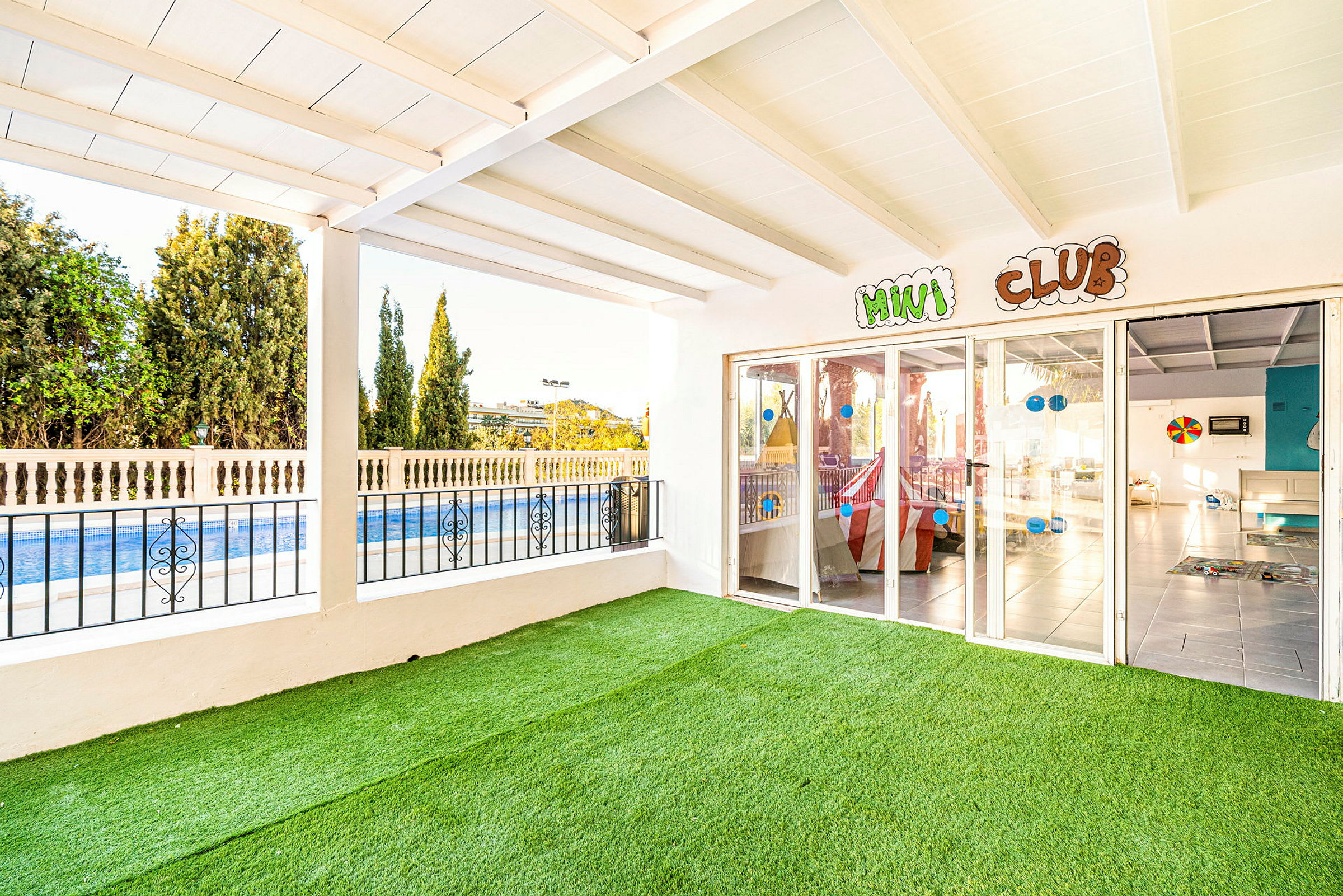 Bild på en utomhusterrass med konstgräs, utsikt över en pool och glasväggar till ett aktivitetsrum märkt 'Mini Club'. (AI-genererad text)