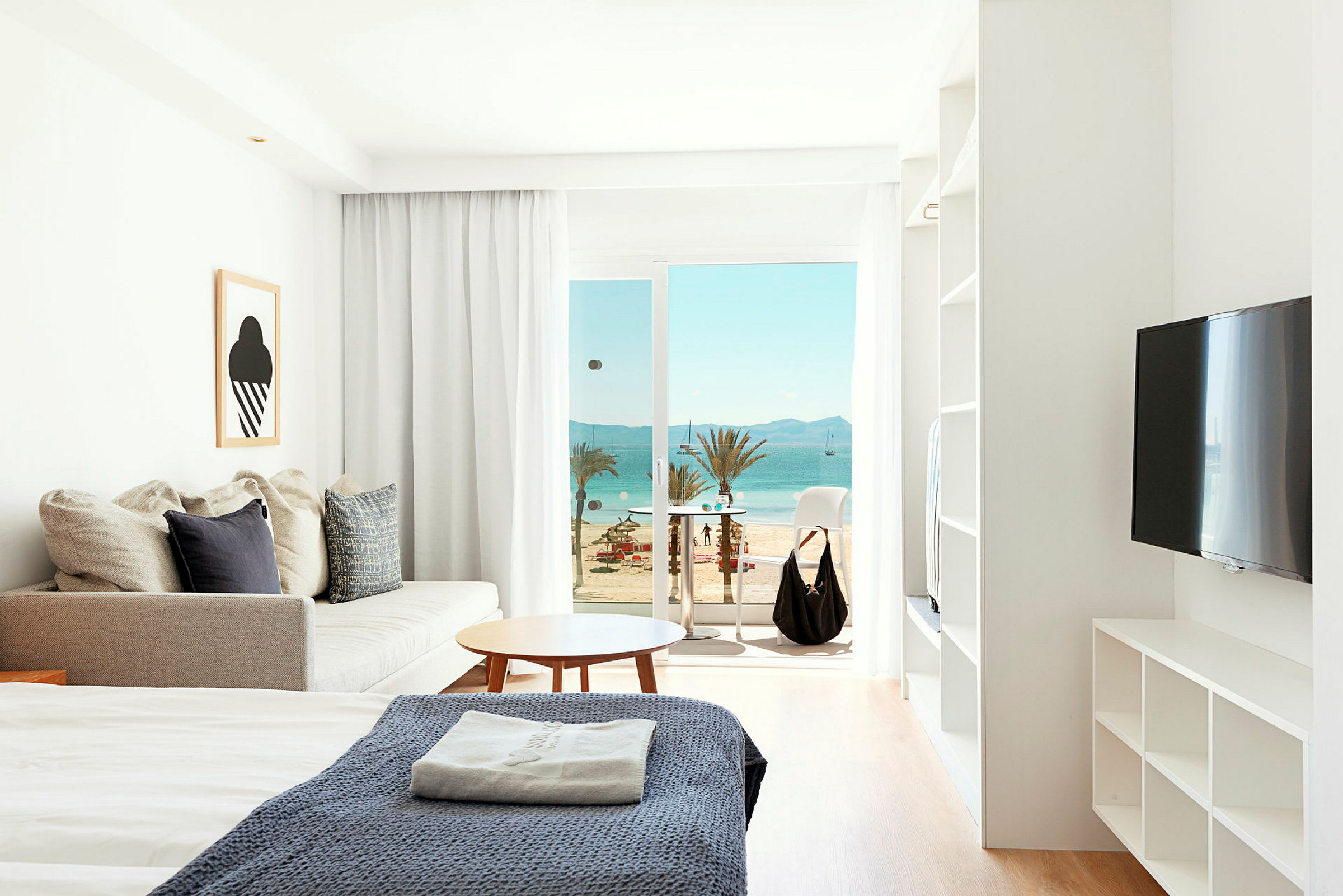 Ett hotellrum med säng, soffa och utsikt över stranden genom stora glasdörrar. (AI-genererad text)