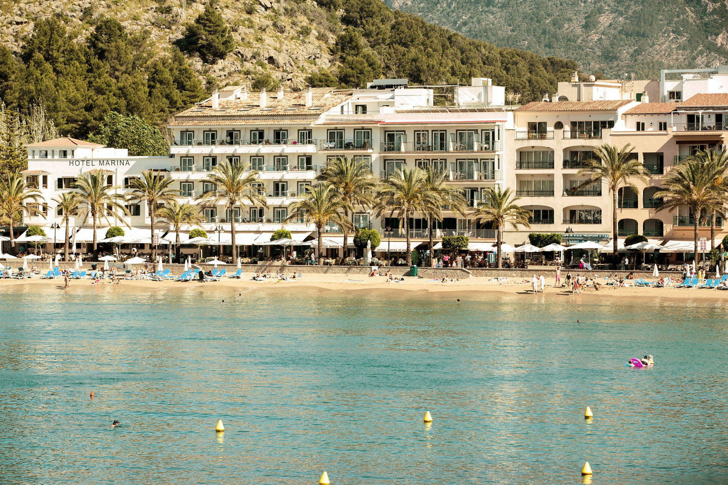 Hotell och strand vid en kust med berg och träd i bakgrunden. (AI-genererad text)