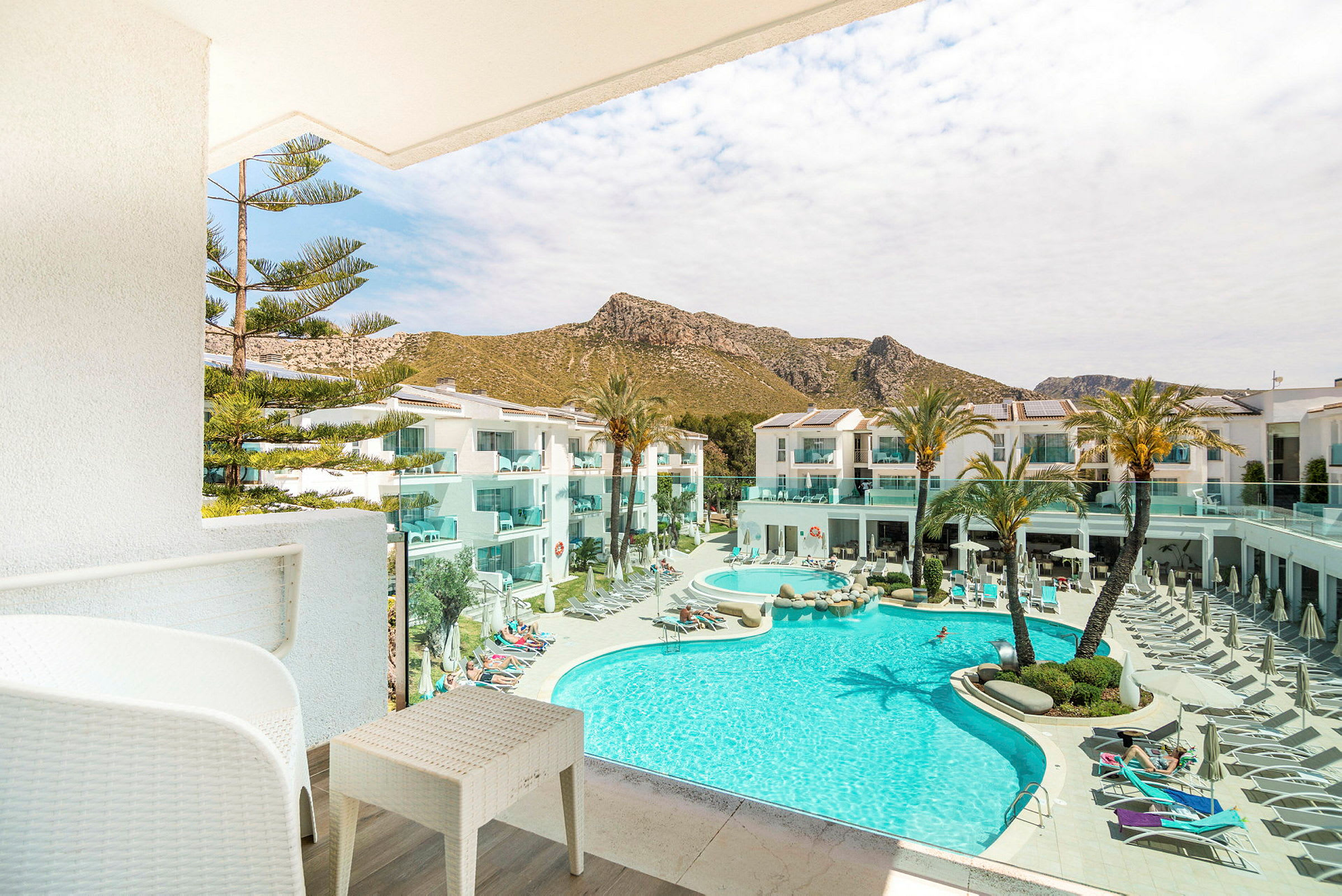 Bilden visar en balkong med utsikt över en pool och hotellbyggnader, omgivna av palmer och berg. (AI-genererad text)