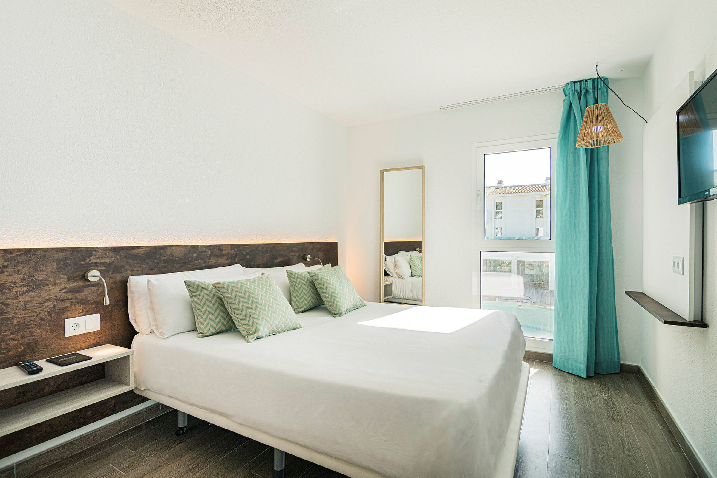 Ett hotellrum med en dubbelsäng, dekorativa kuddar, ett fönster med turkosa gardiner och en tv på väggen. (AI-genererad text)