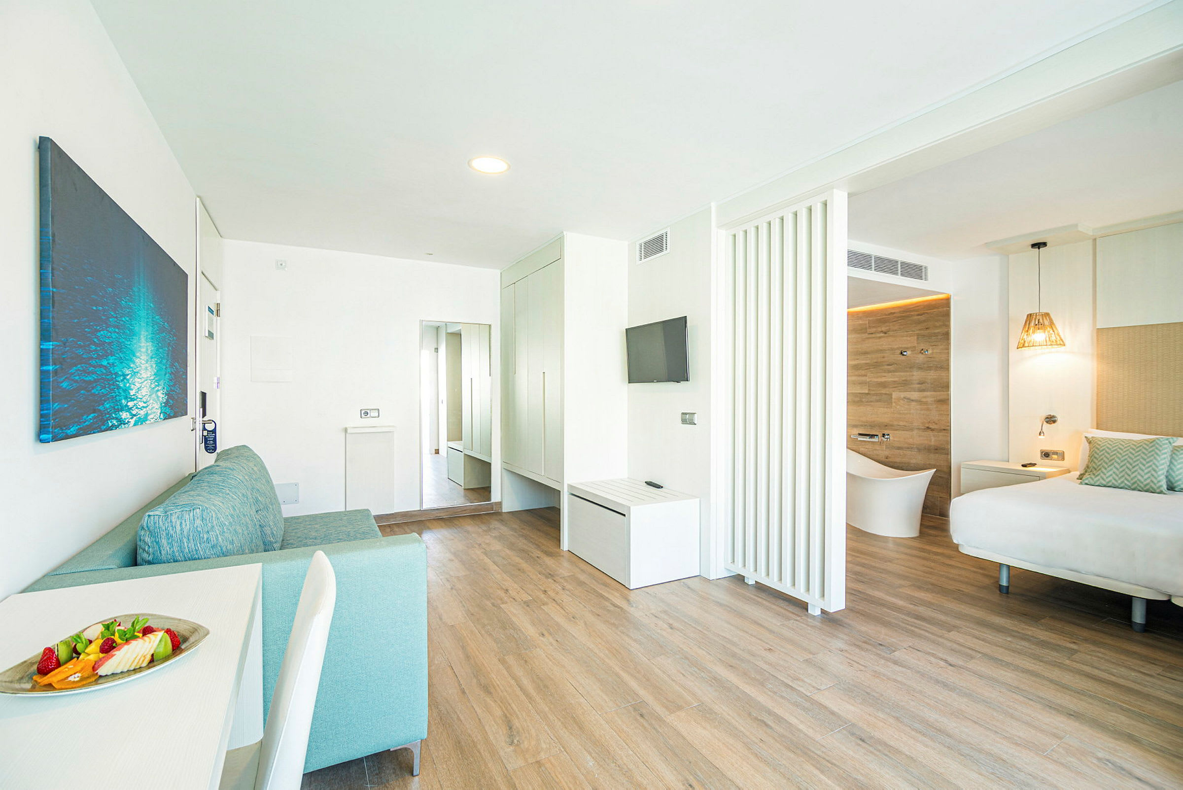 Ett modernt hotellrum med ljus inredning, trägolv och separata sov- och vardagsrumsområden. En tallrik med frukt är placerad på bordet. (AI-genererad text)