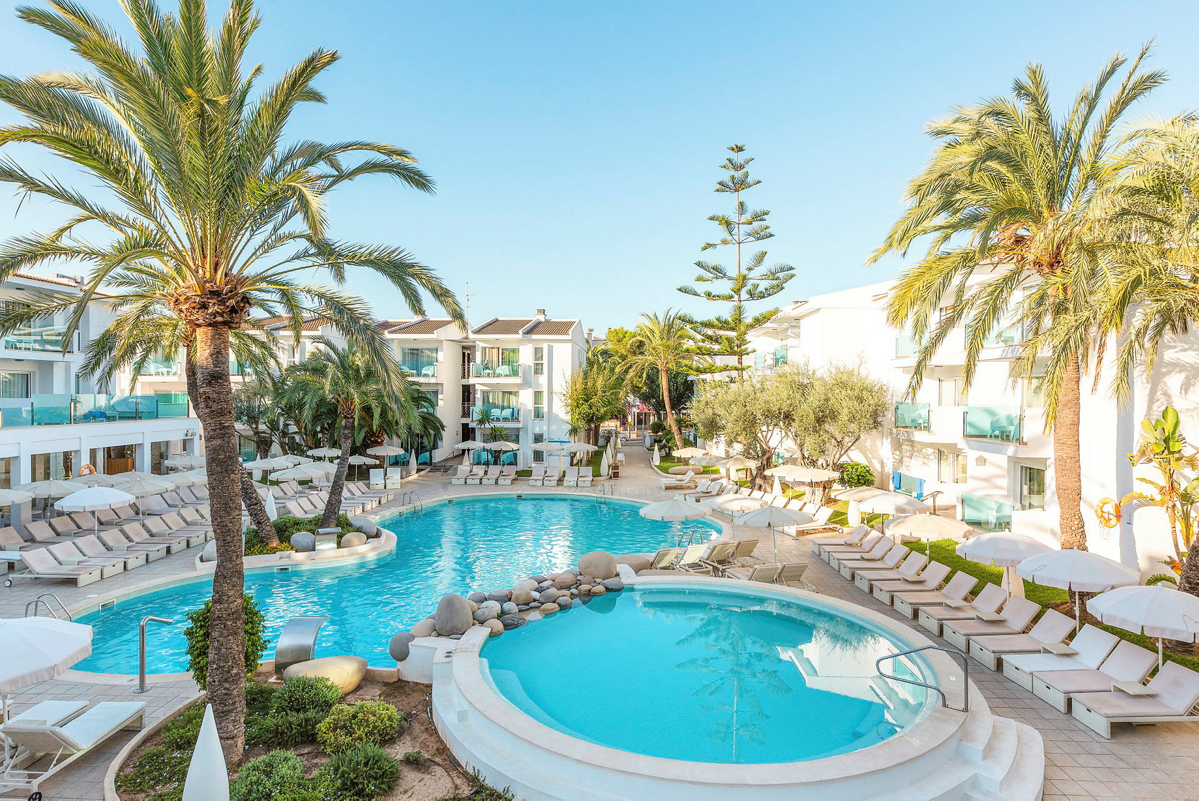 Bild av hotellområde med två pooler omgivet av solstolar och palmträd. (AI-genererad text)