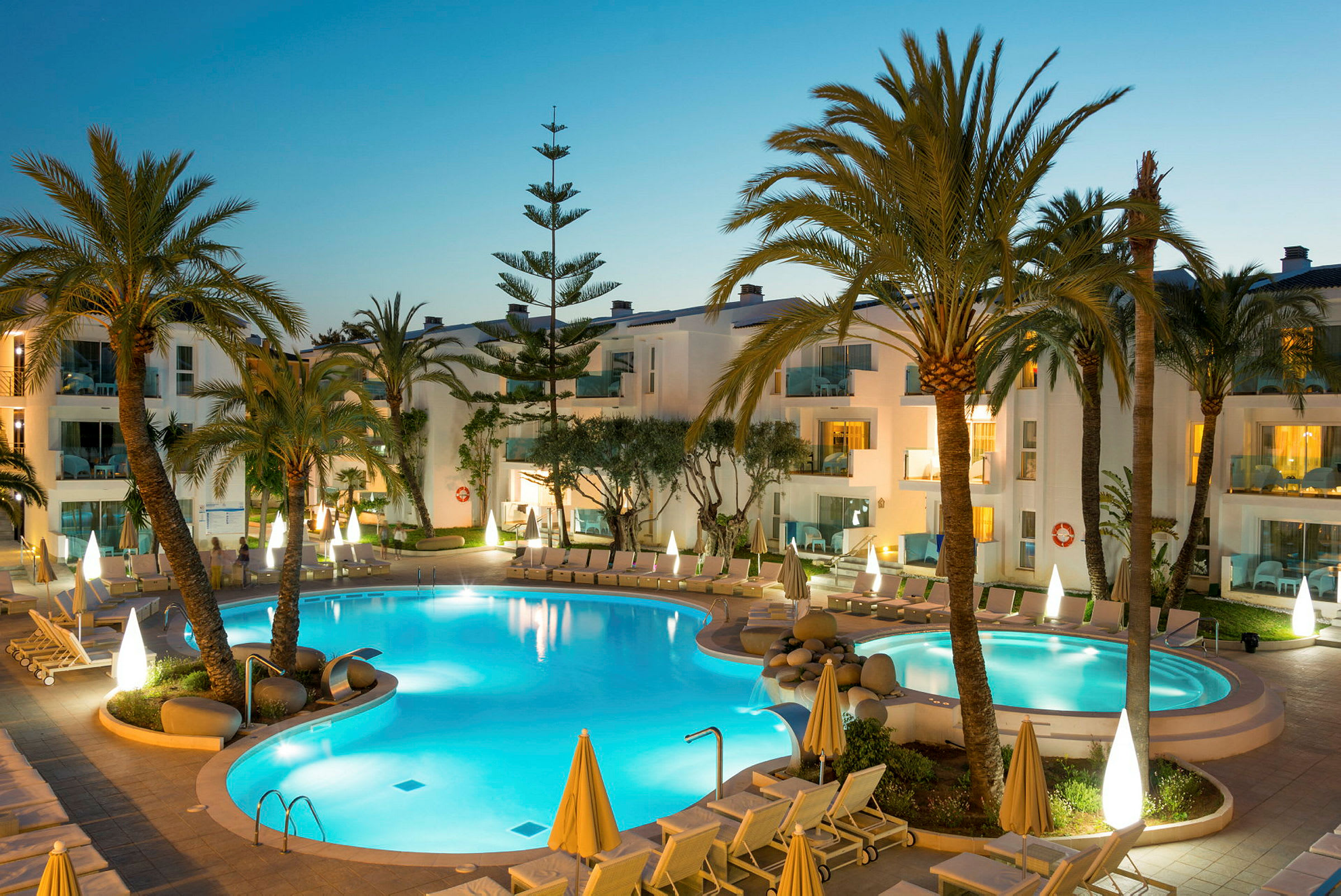 En upplyst pool omgiven av solstolar och palmer vid ett hotellkomplex. (AI-genererad text)