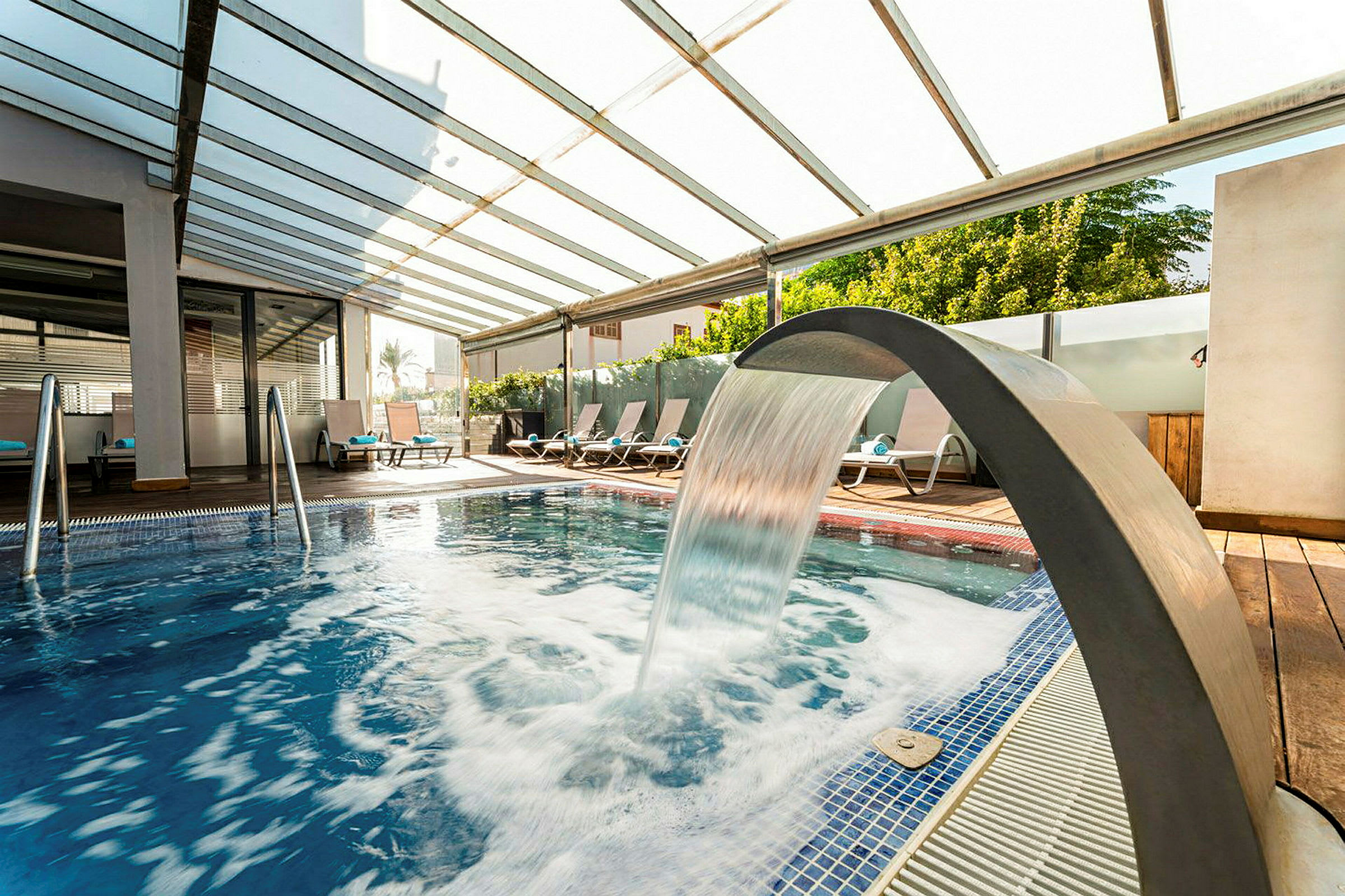 En pool med solstolar och ett vattenfall under ett delvis öppet tak. (AI-genererad text)