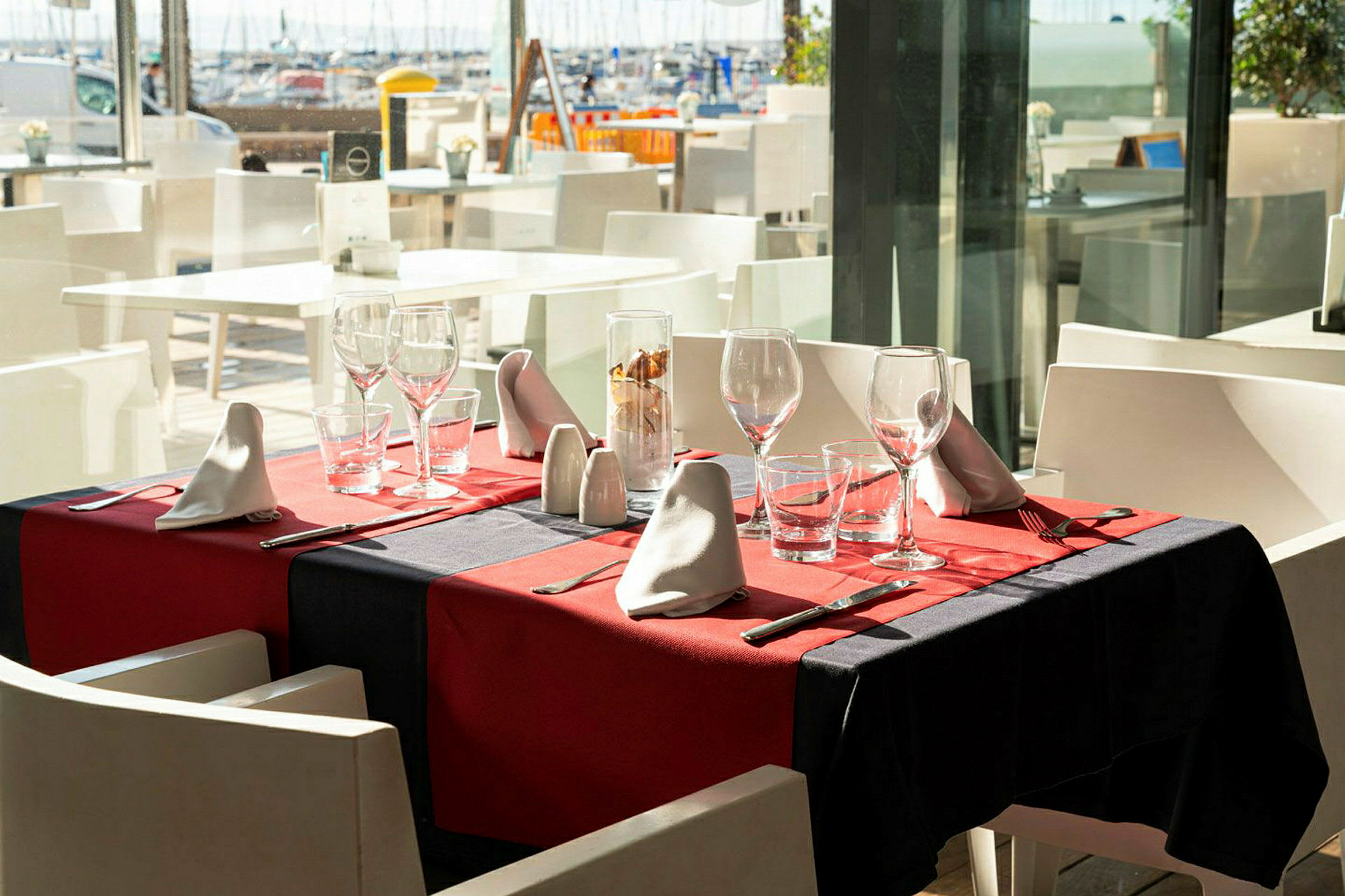Ett dukat bord med röd duk, glas och bestick på en restaurang med utsikt över hamnen. (AI-genererad text)