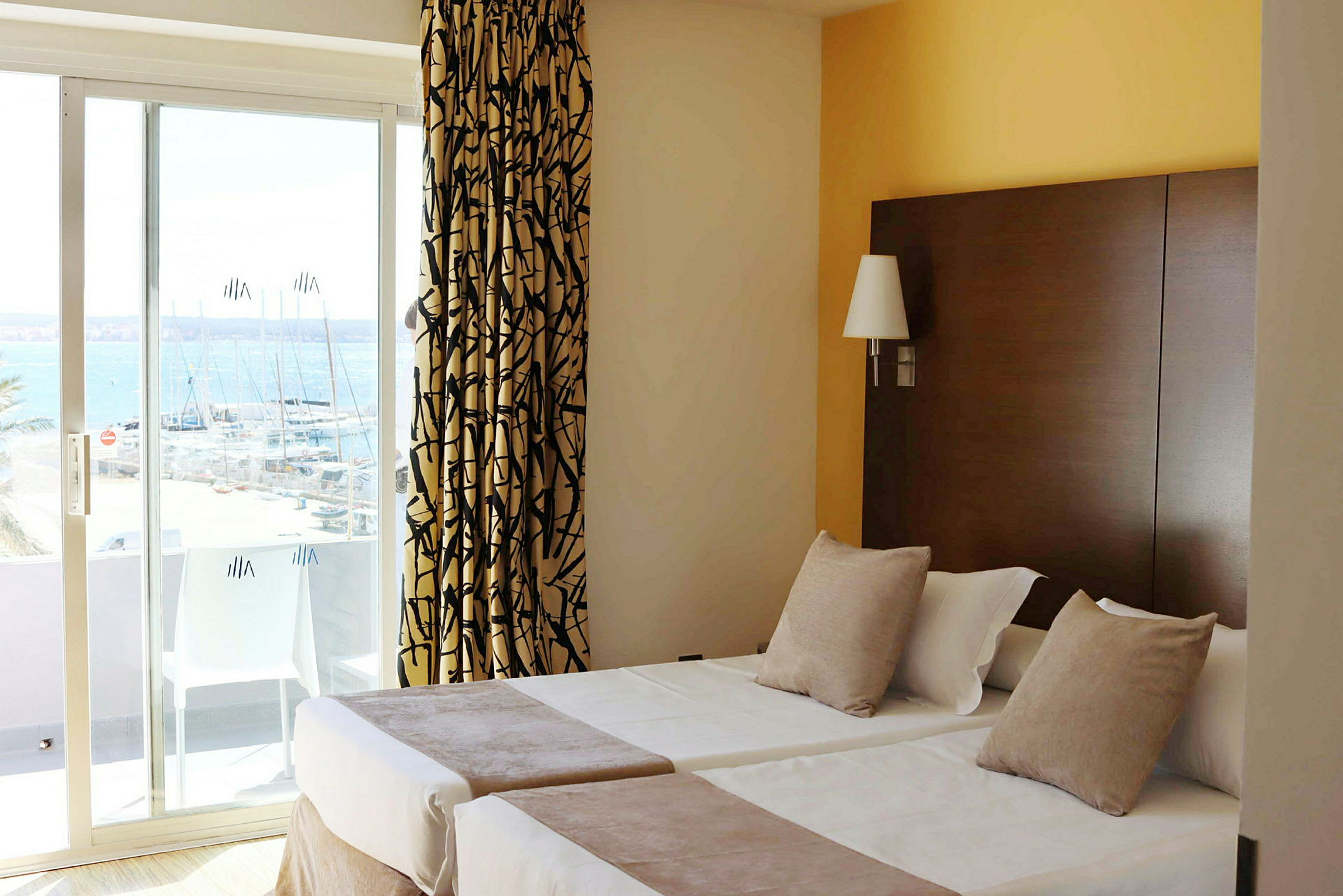 Hotellrum med två enkelsängar, dekorerade med kuddar och överkast. Ett fönster med skjutdörr visar utsikt över en marina. (AI-genererad text)