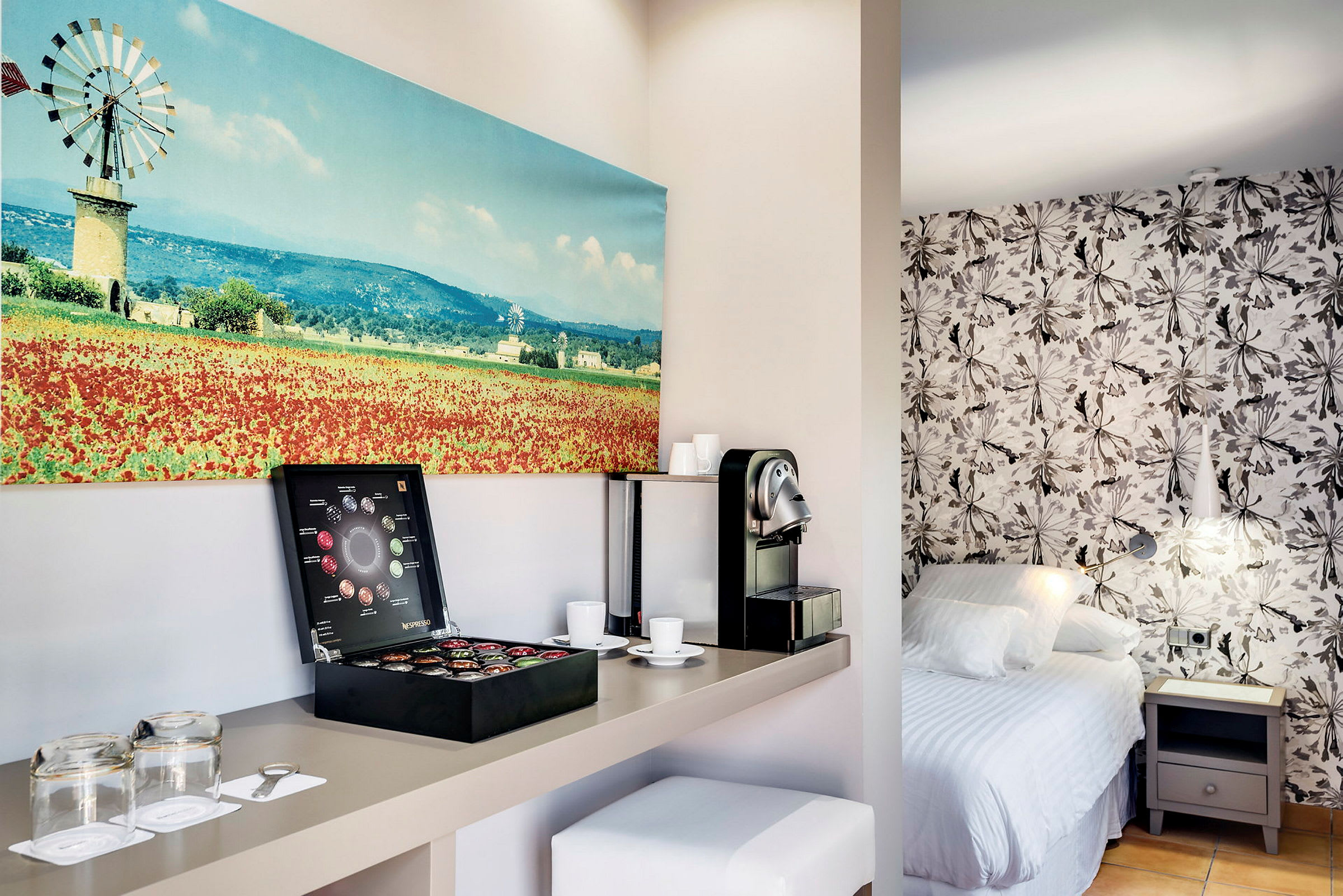Ett hotellrum med en kaffemaskin och kaffekapslar på en hylla, en säng med blommigt mönstrad vägg och en målning av en landskapsutsikt. (AI-genererad text)