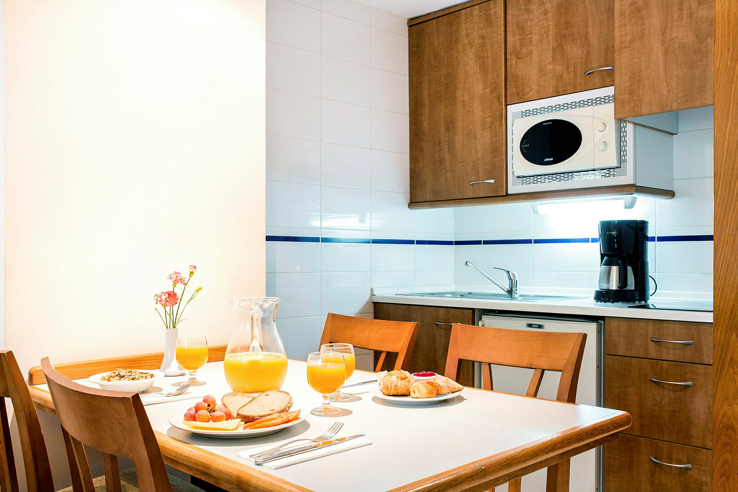 En bild av ett kök med ett träbord dukat för frukost samt en diskho och köksskåp i bakgrunden. (AI-genererad text)