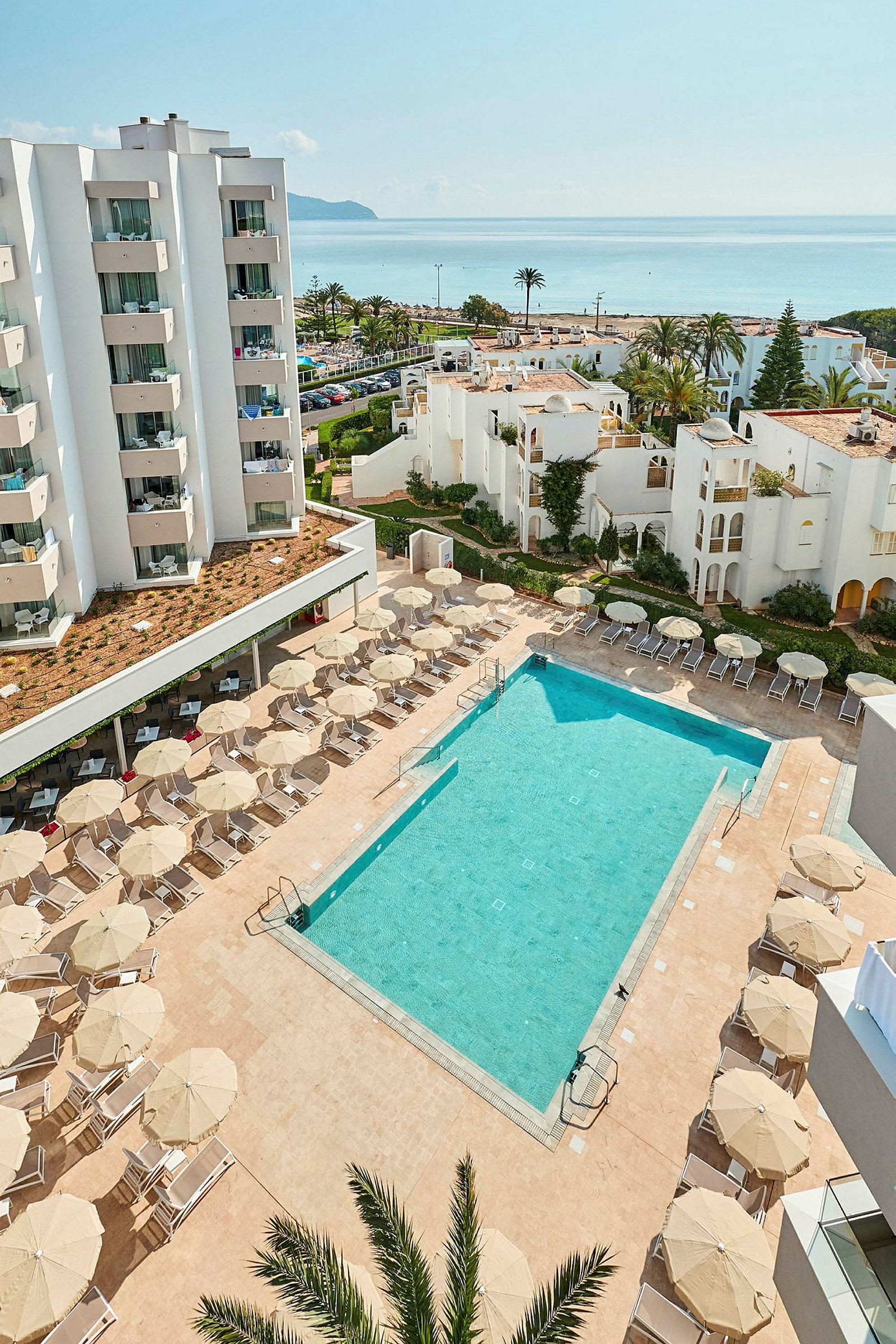 Bilden visar en utomhuspool framför ett hotell med solstolar och parasoller samt vitkalkade byggnader och havet i bakgrunden. (AI-genererad text)