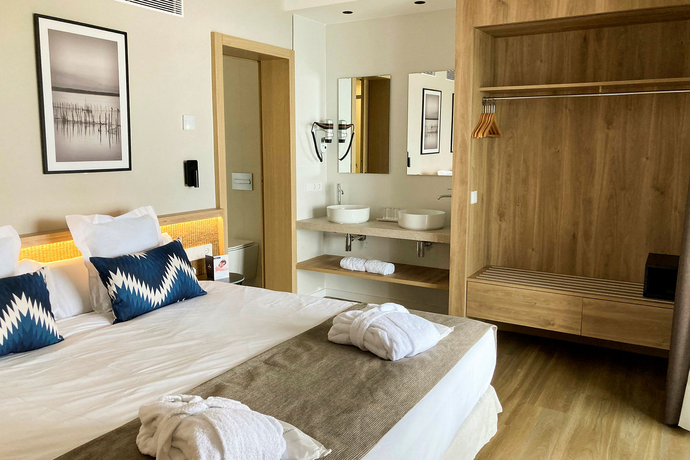 Ett hotellrum med en dubbelsäng, två blå kuddar och vita badrockar på sängen. I bakgrunden syns ett badrum med två handfat och en spegel samt en garderob med träpanel. (AI-genererad text)