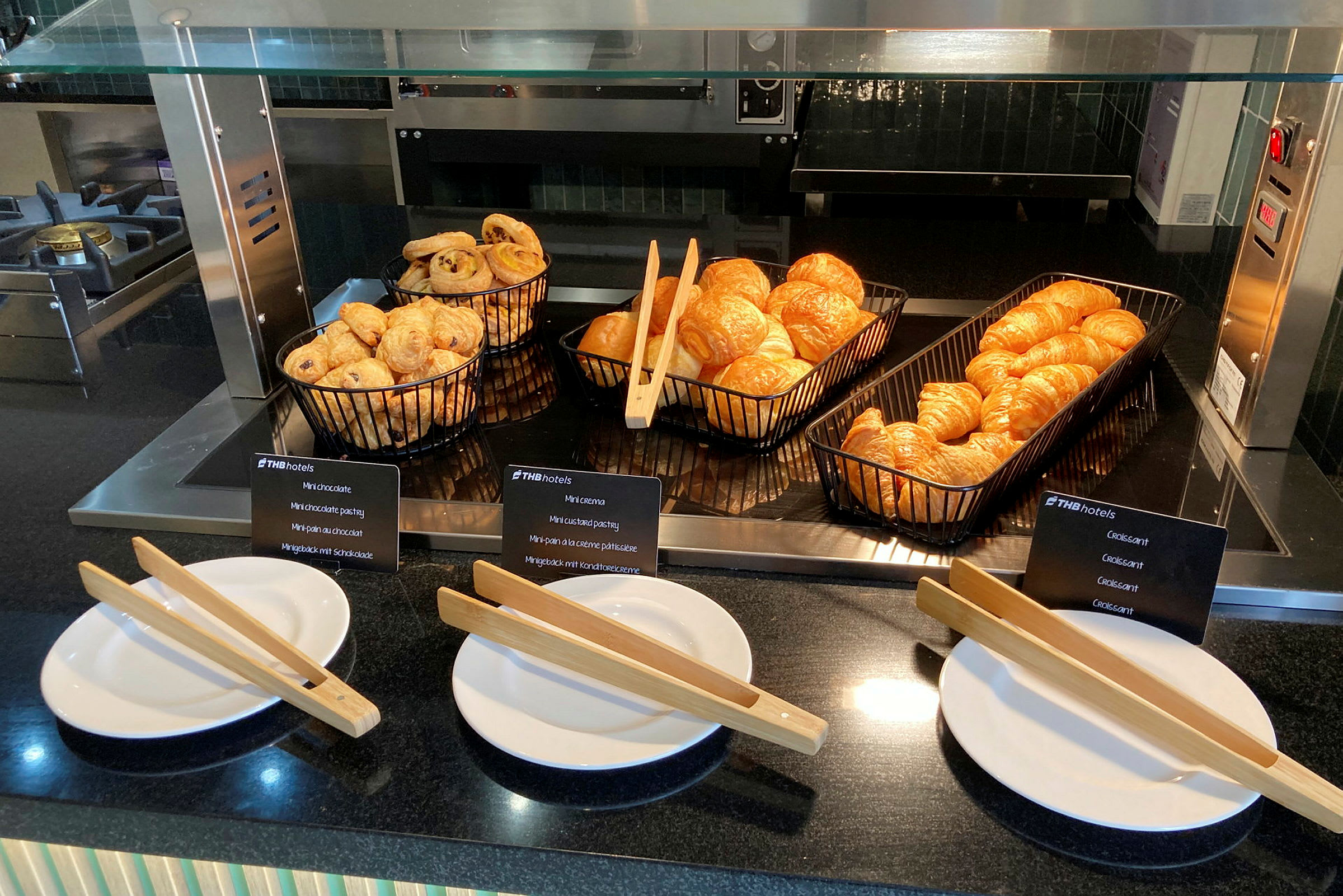 Frukostbuffé med nygräddade croissanter och bakverk i svarta korgar. Framför ligger vita tallrikar med träpincetter. (AI-genererad text)