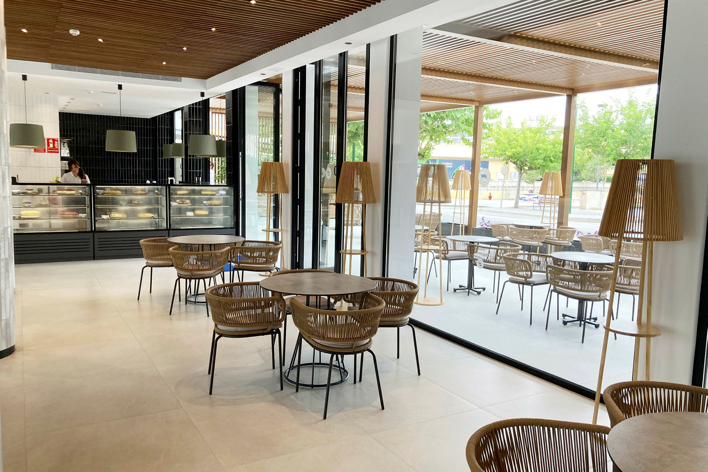 Ett café med moderna möbler, stora fönster och glasväggar finns på bilden. Det finns flera bord och stolar samt en disk med bakverk. (AI-genererad text)