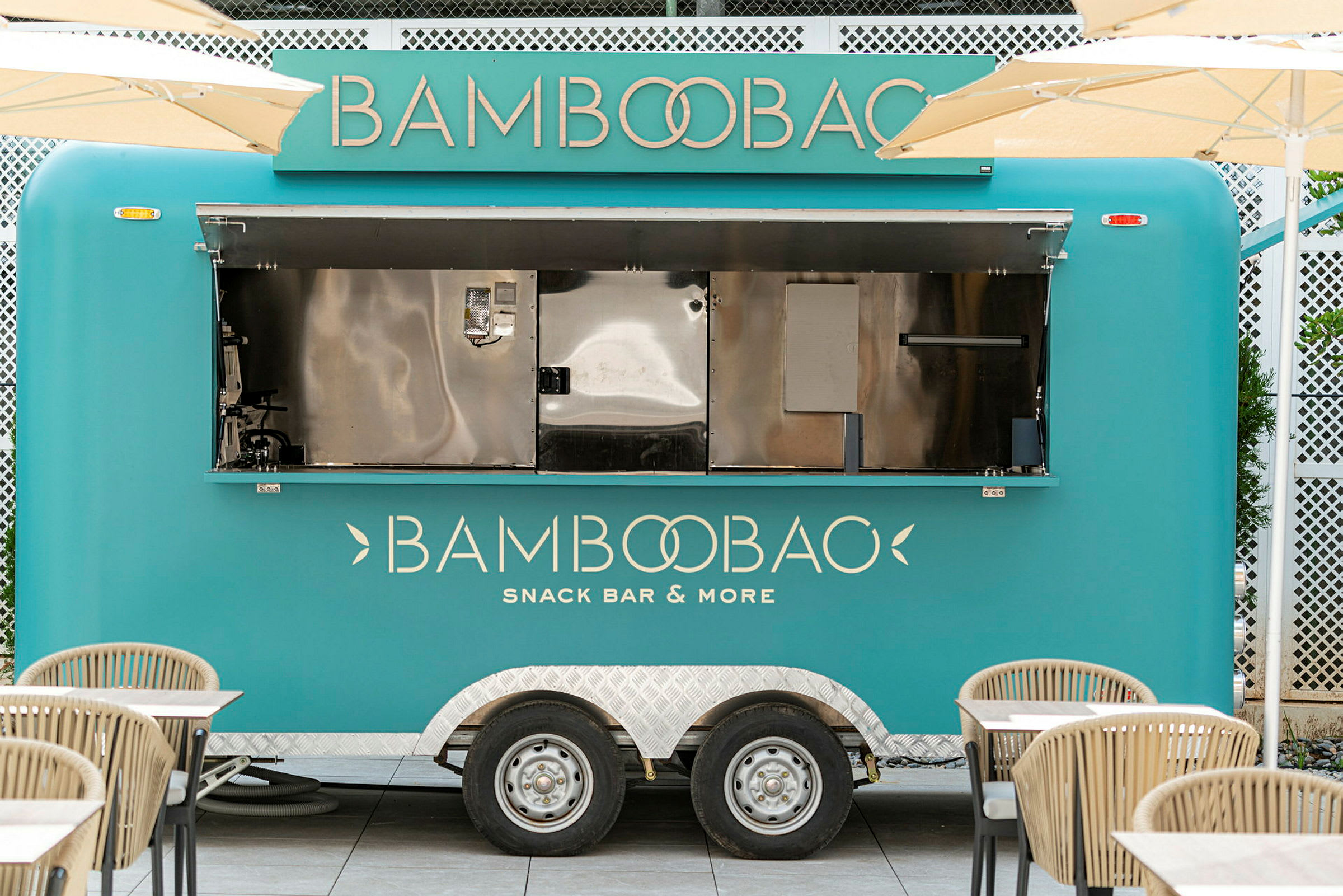 En turkos matvagn med texten 'Bamboobao Snack Bar & More' står parkerad omgiven av bord och stolar. (AI-genererad text)