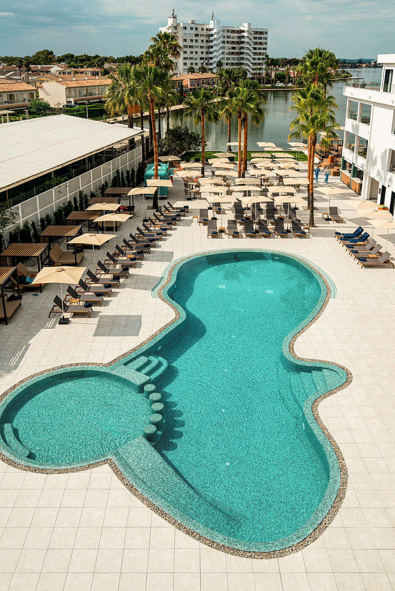 En pool med solstolar och parasoller runt omkring på en terrass vid ett hotell. Byggnader och en sjö syns i bakgrunden. (AI-genererad text)