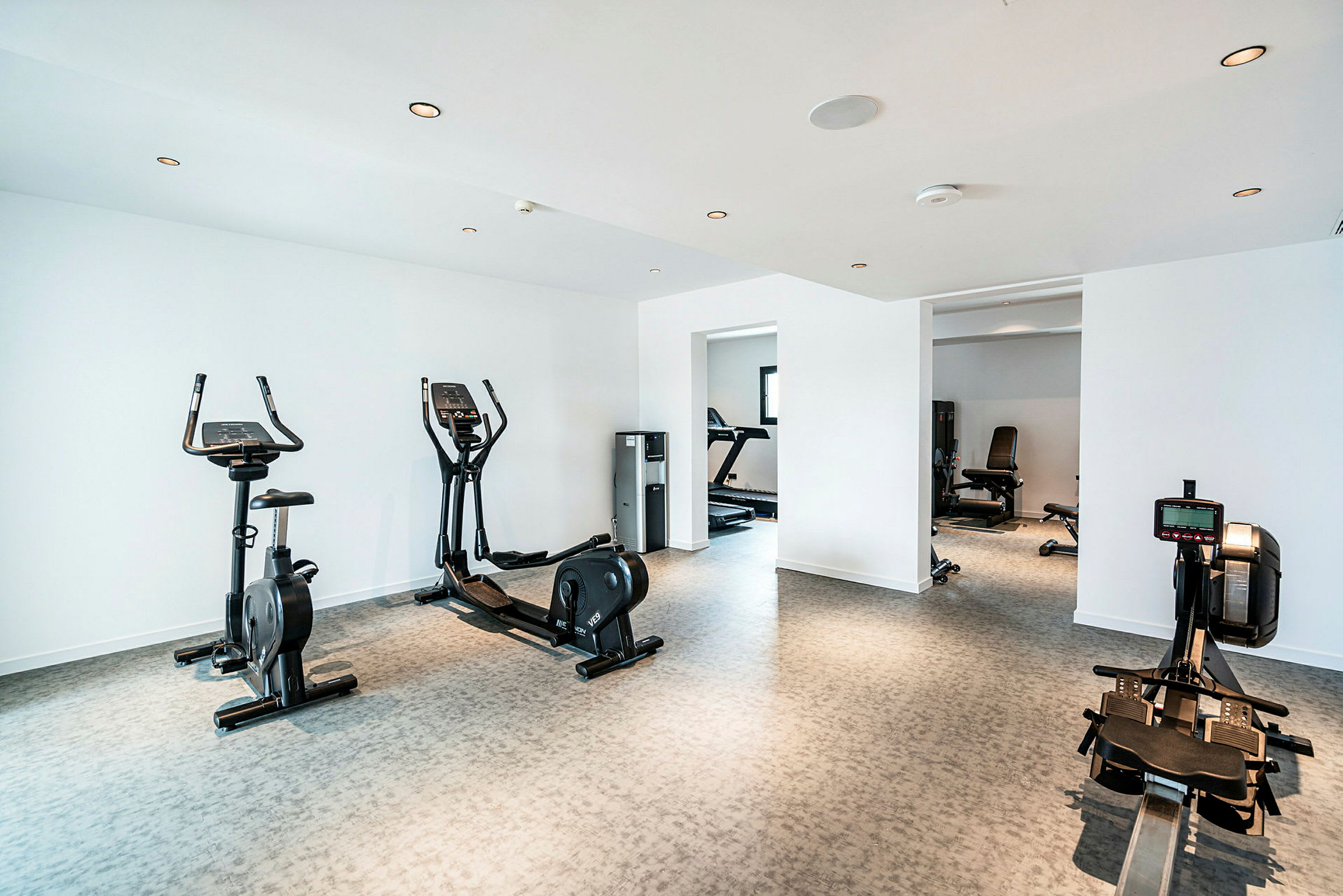 Ett ljust gymrum med träningsutrustning såsom motionscykel, roddmaskin och crosstrainer samt fönster och andra träningsmaskiner i bakgrunden. (AI-genererad text)