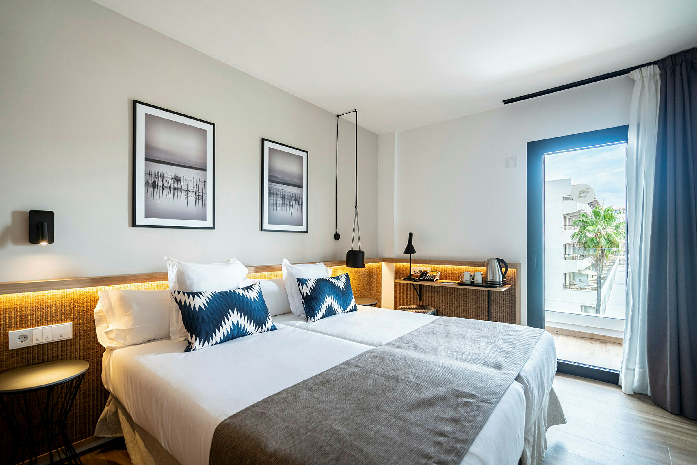 Ett hotellrum med två sängar täckta av vita lakan och grå överkast. Det finns blå dekorativa kuddar på sängarna. Väggarna har två inramade konstbilder, och ett fönster visar utsikt över en palm och byggnader. (AI-genererad text)