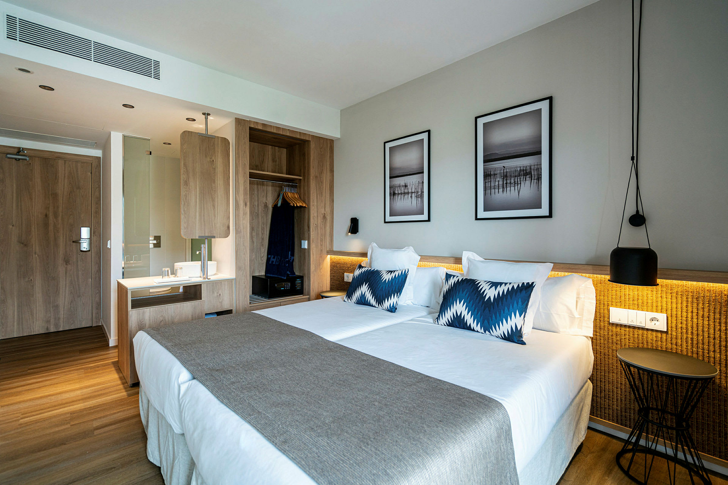 Ett hotellrum med en stor dubbelsäng, beige överkast och blåvita kuddar. Väggen har två inramade tavlor ovanför sängen, och till vänster finns en träpanel med garderob och skrivbord. (AI-genererad text)