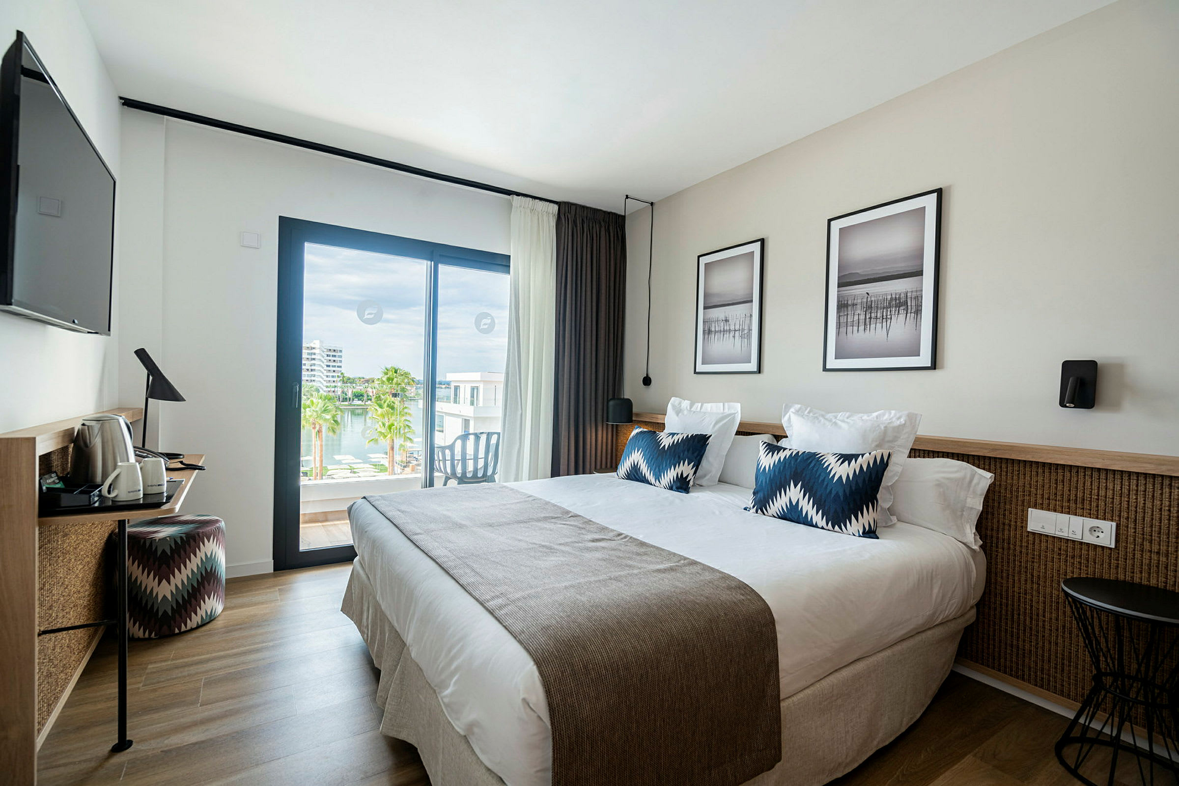 Ett modernt hotellrum med en dubbelsäng, två kuddar och en filt, samt väggkonst ovanför sängen. Rummet har en skrivbordshörna med en stol och en tv på väggen. En stor glasdörr leder ut till en balkong med utsikt över vatten och palmer. (AI-genererad text)