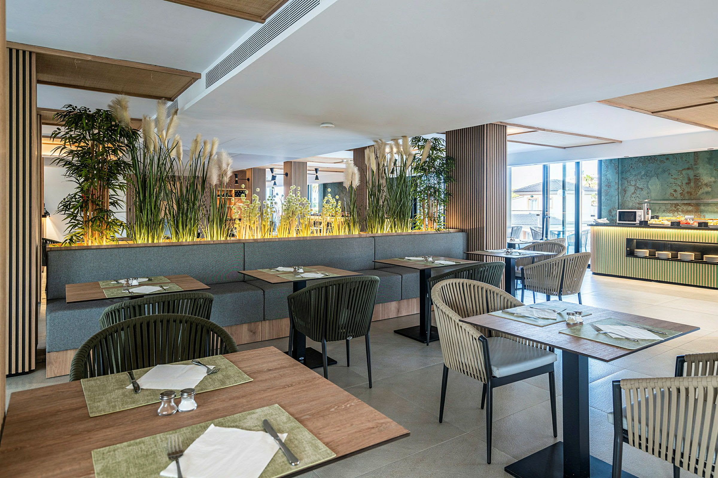 Bild av en modern restaurang med sittplatser, bord och växter som dekoration. Fönster släpper in naturligt ljus. (AI-genererad text)