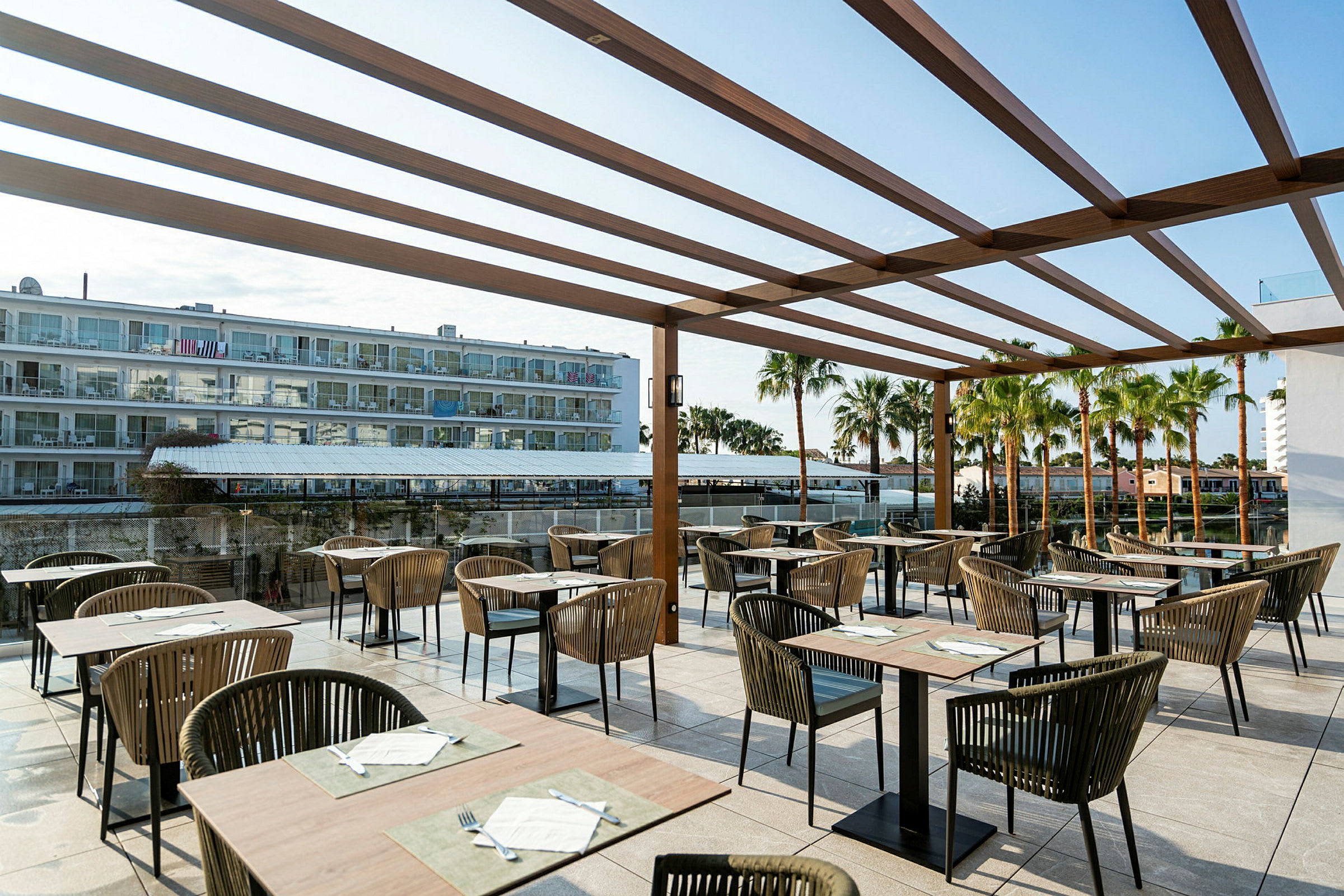 Bild av en restaurangterrass med bord och stolar under en träpergola. I bakgrunden syns ett hotell och palmer. (AI-genererad text)