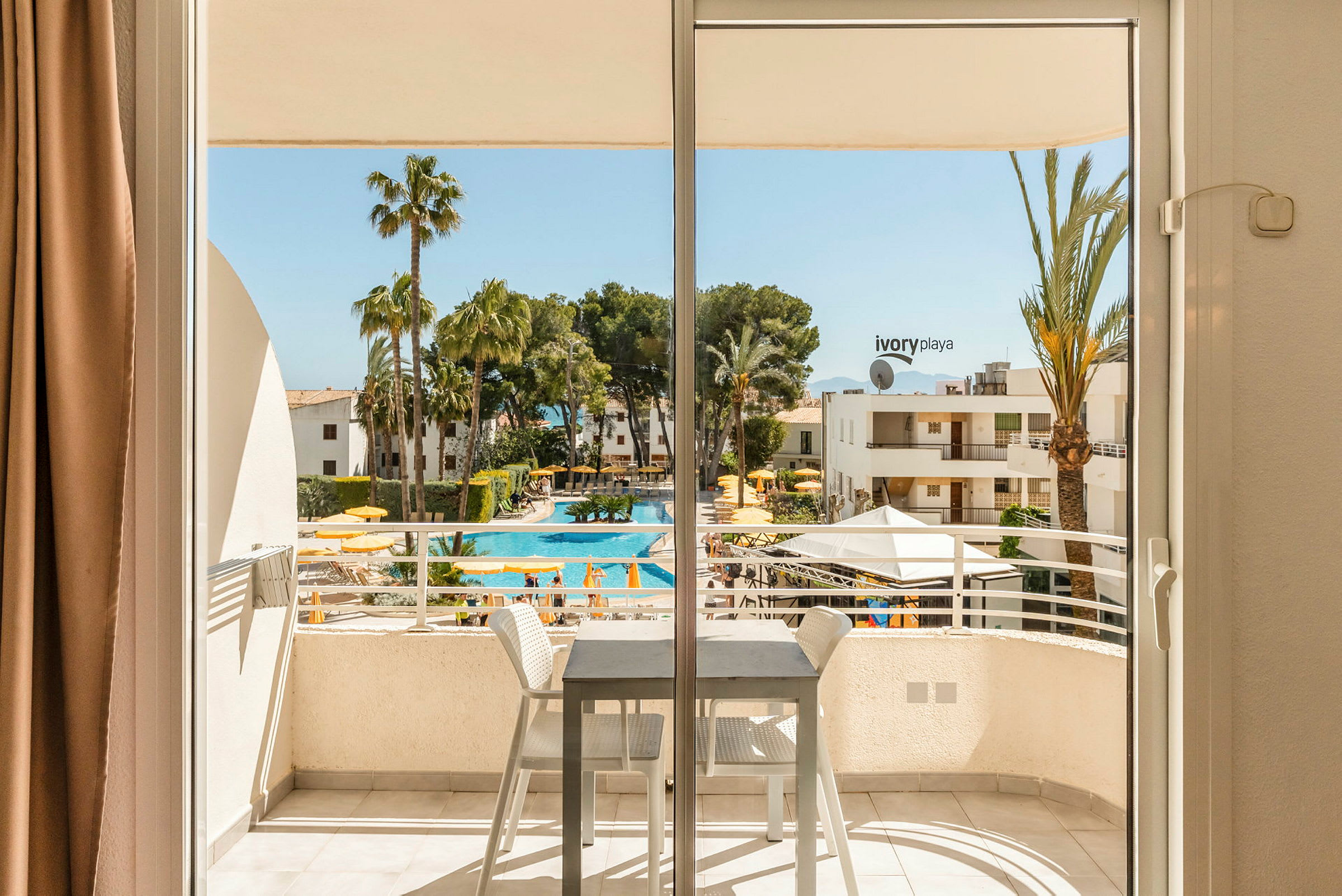 Vy från ett hotellrum med balkong där utsikten visar en pool omgiven av gula parasoller och palmer. (AI-genererad text)
