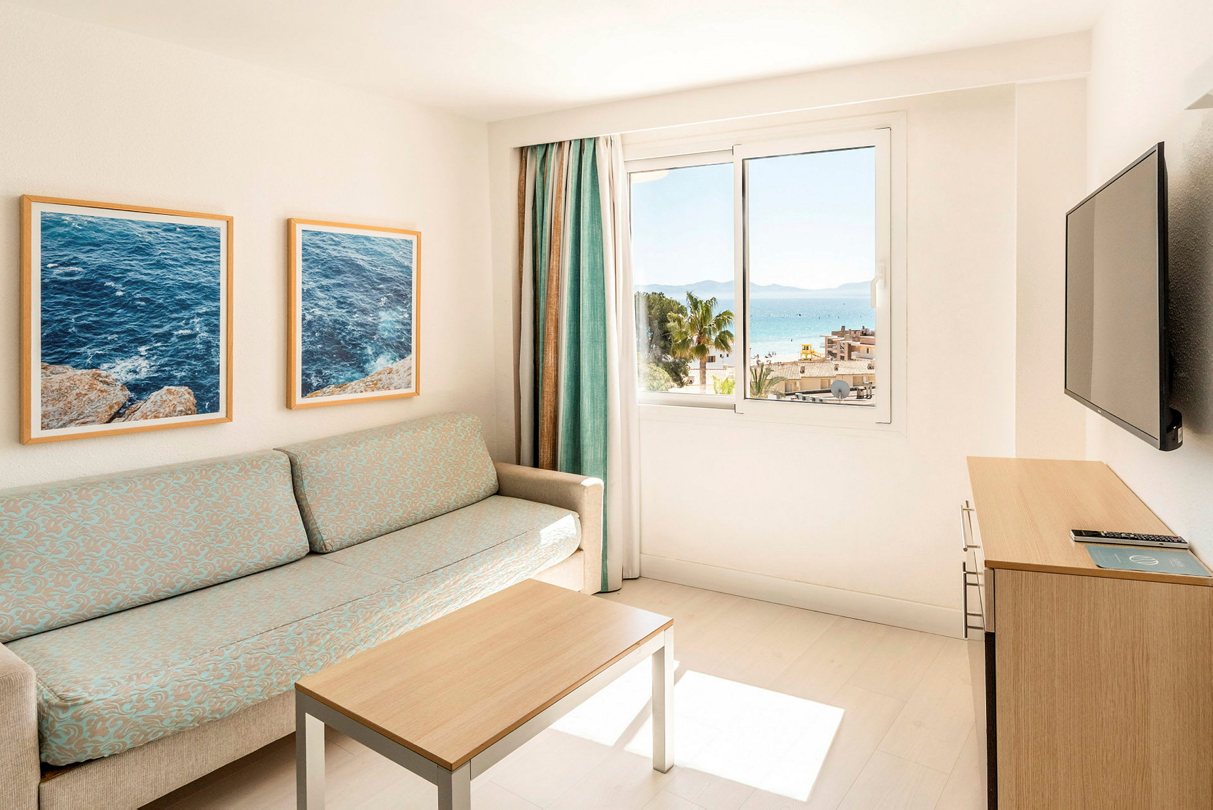 Ett ljust hotellrum med en soffa, bord, och en TV. Fönstret visar en utsikt över havet och palmer. (AI-genererad text)