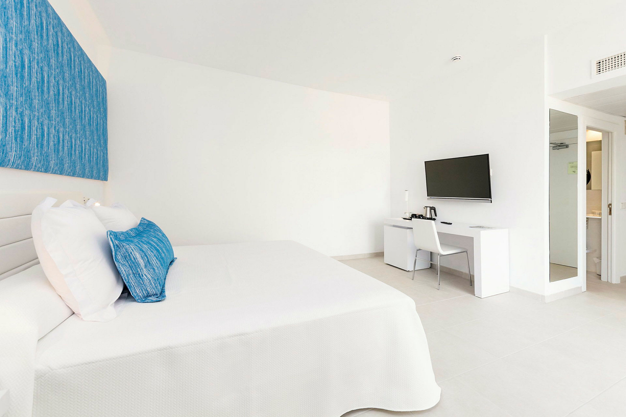 Ett hotellrum med en vit säng klädd med blå kuddar och ett angränsande skrivbord med en väggmonterad TV ovanför. (AI-genererad text)