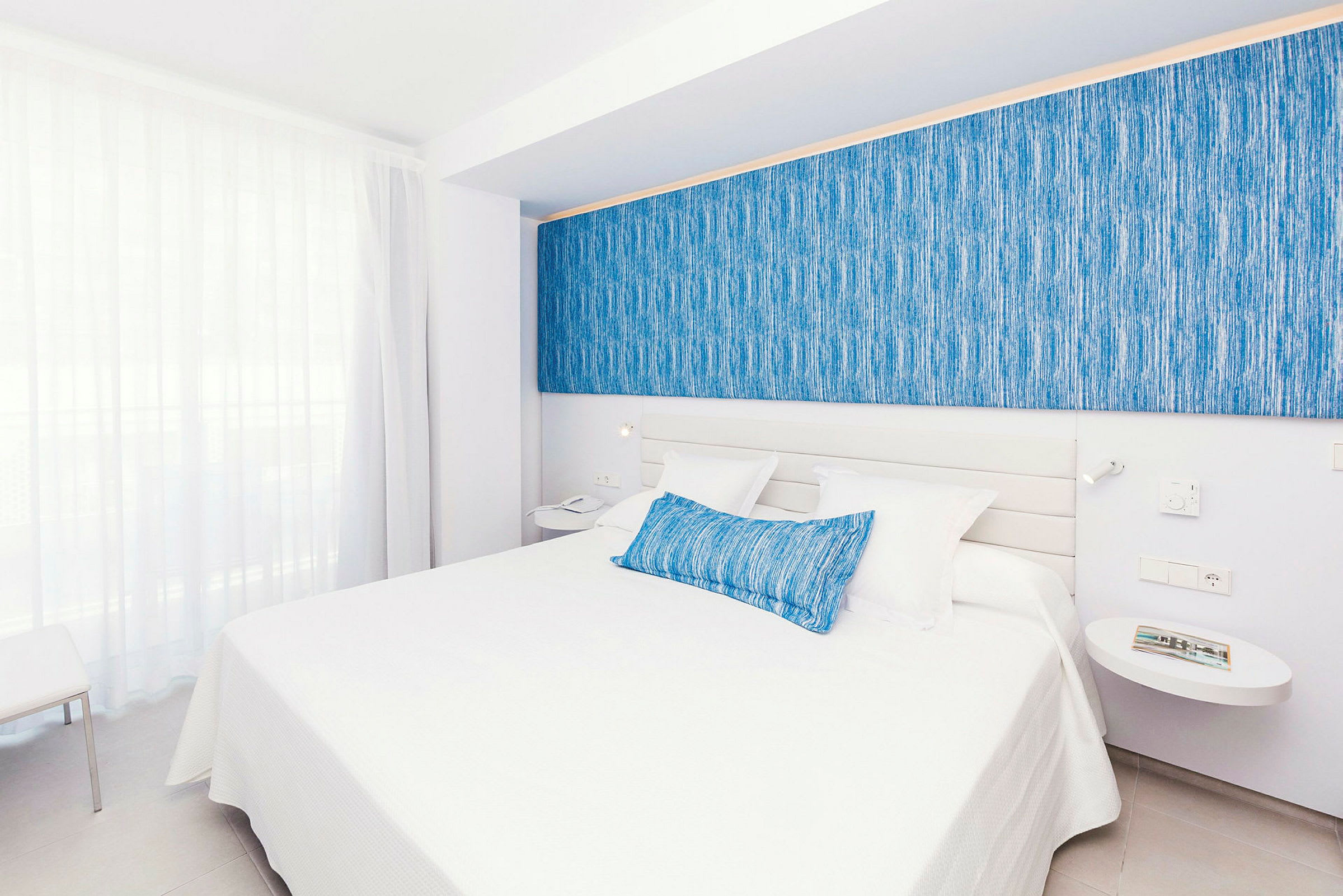 Ett hotellrum med en dubbelsäng, vita sängkläder och blåa dekorativa inslag. (AI-genererad text)