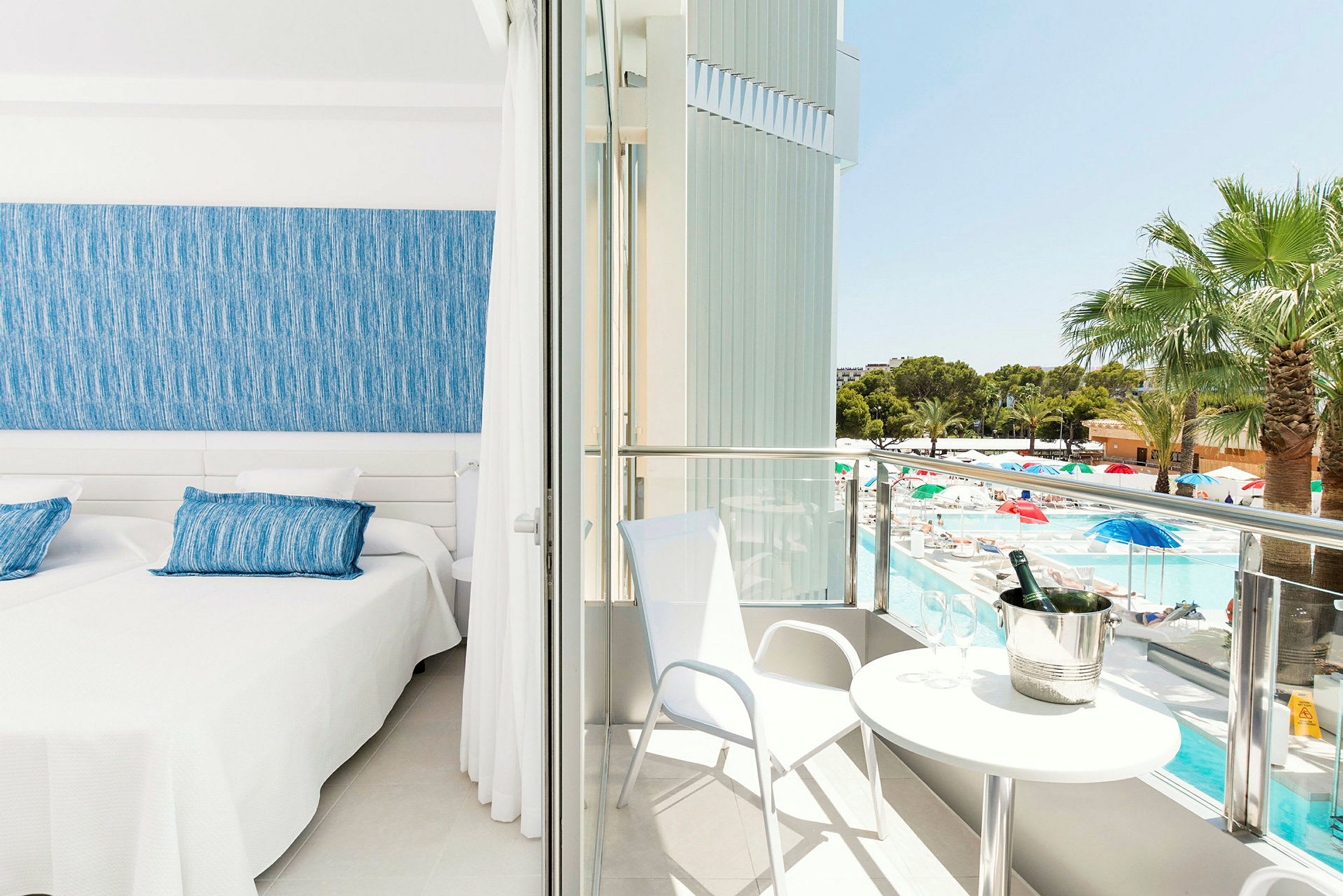 Ett hotellrum med två sängar, en blå sänggavel och tillgång till en balkong med utsikt över en pool och trädgård. (AI-genererad text)