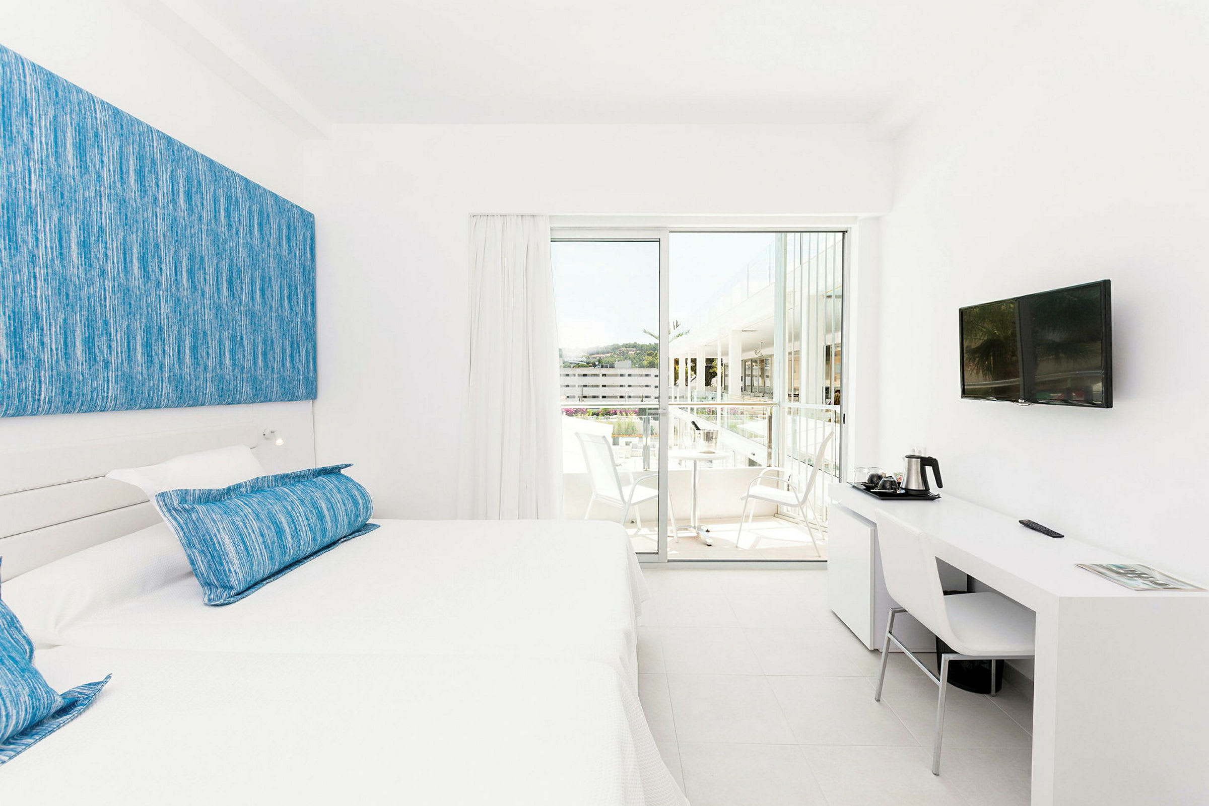 Ett ljust hotellrum med en vit inredning, två enkelsängar med blå kuddar och ett skrivbord med stol och tv på väggen. En skjutdörr leder ut till balkongen med sittmöbler. (AI-genererad text)
