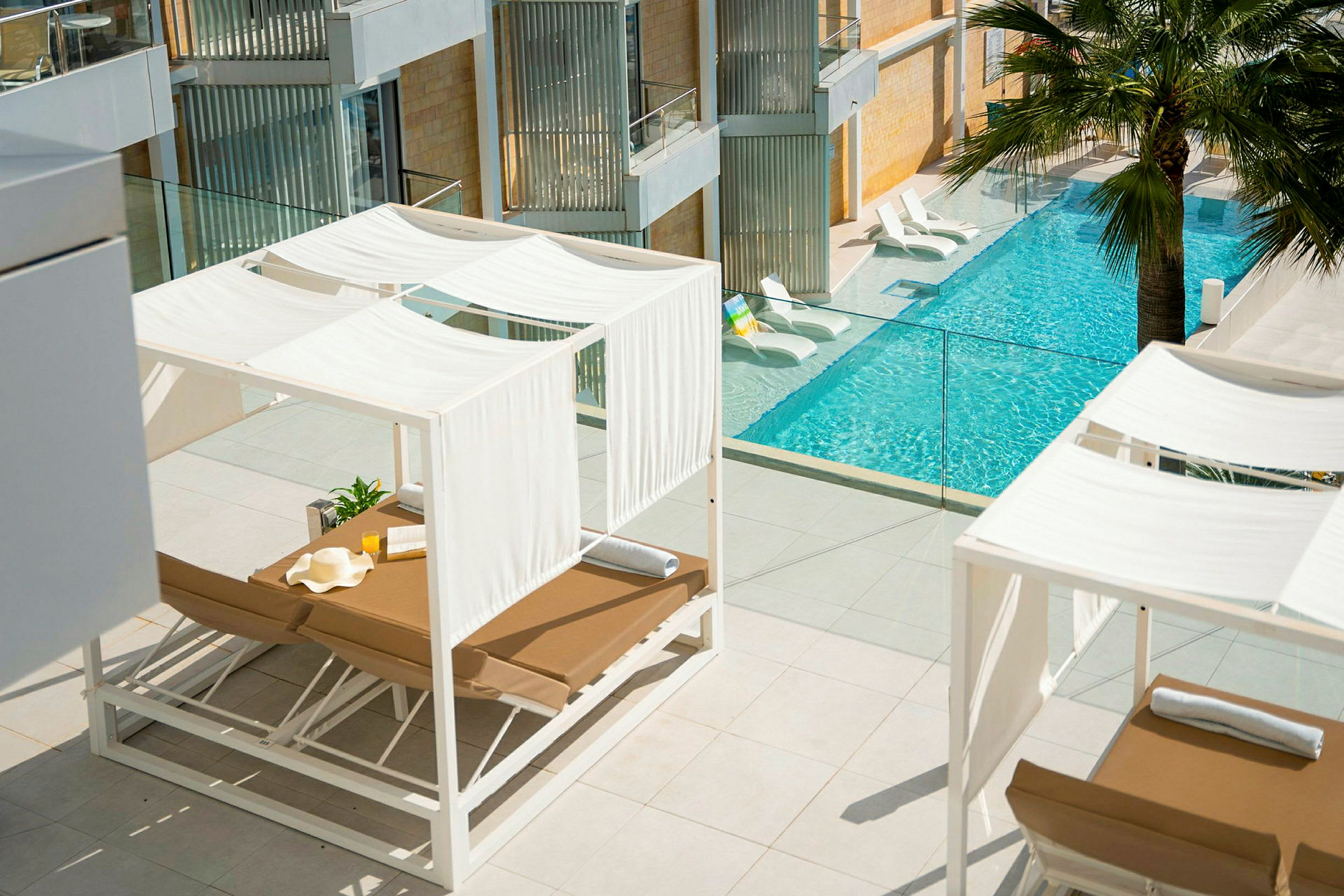 En terrass med vita solstolar under skuggande baldakiner och utsikt över en pool med klart vatten. (AI-genererad text)