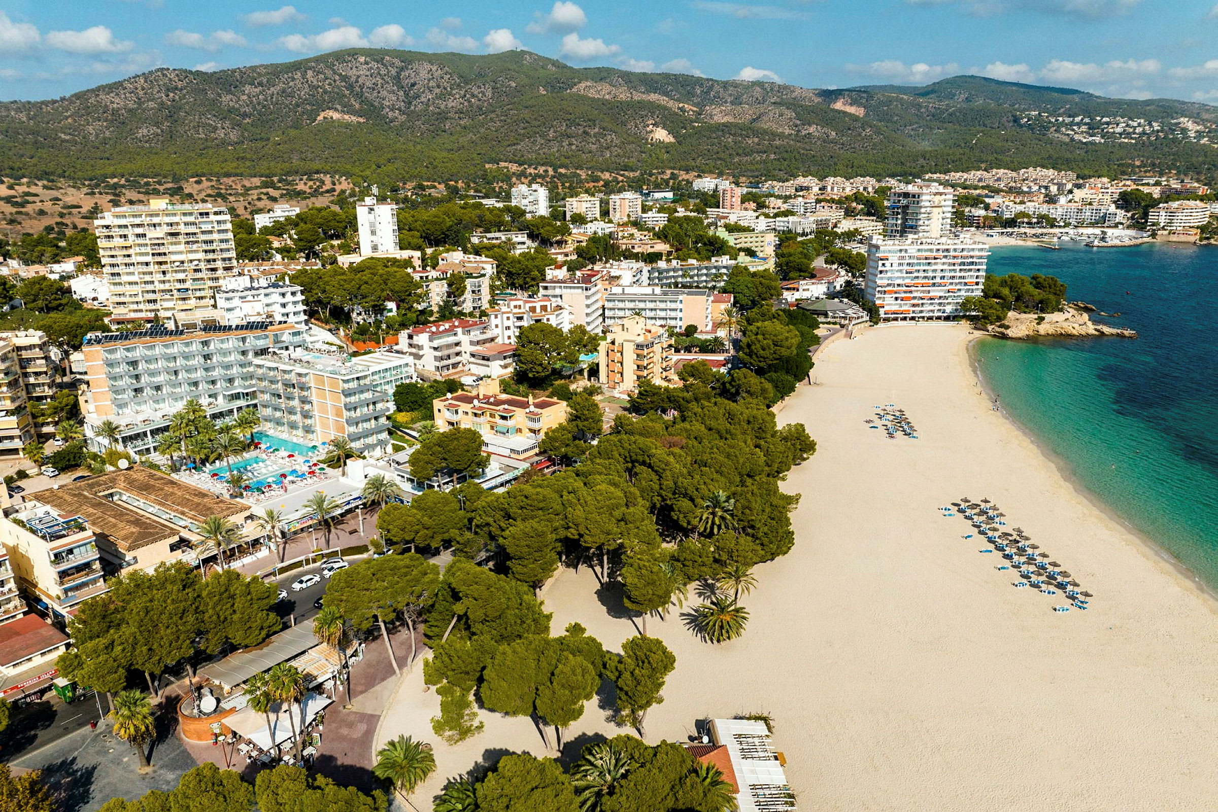 Flygfoto över en sandstrand med parasoller och solstolar, omgiven av träd och hotellbyggnader, med grönskande berg i bakgrunden. (AI-genererad text)