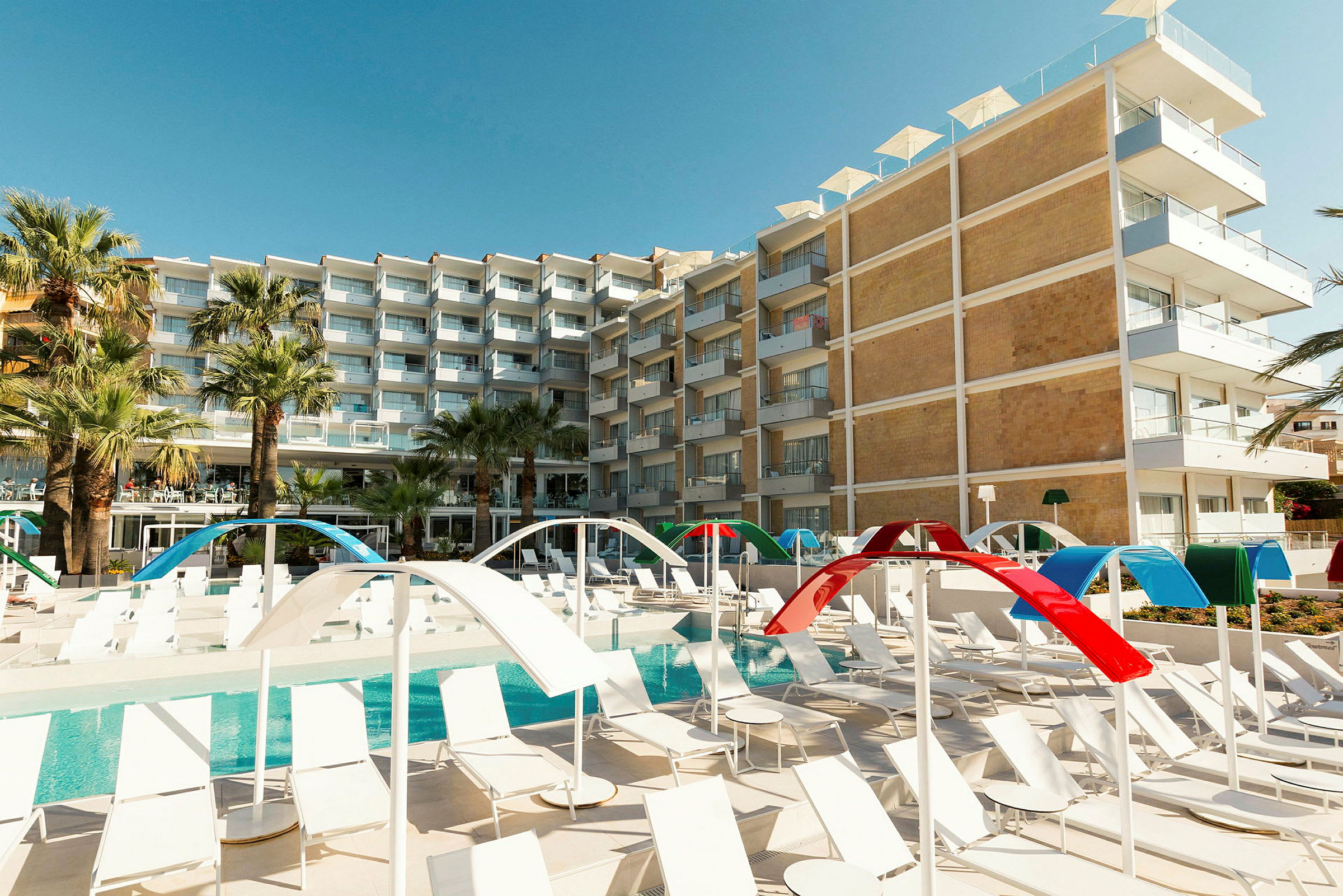 Ett hotell med flera våningar i modern design syns bakom en poolområde med solstolar och färgglada solskydd. (AI-genererad text)