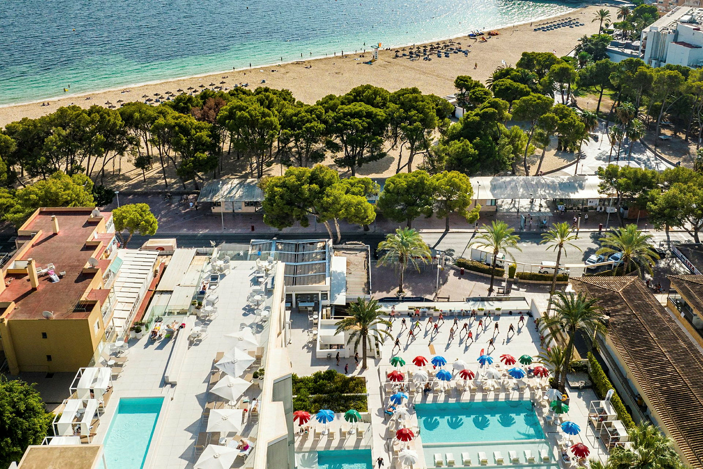 Vy från ovan av ett hotellområde med utomhuspooler, parasoller och solsängar. En sandstrand och hav syns i bakgrunden. (AI-genererad text)