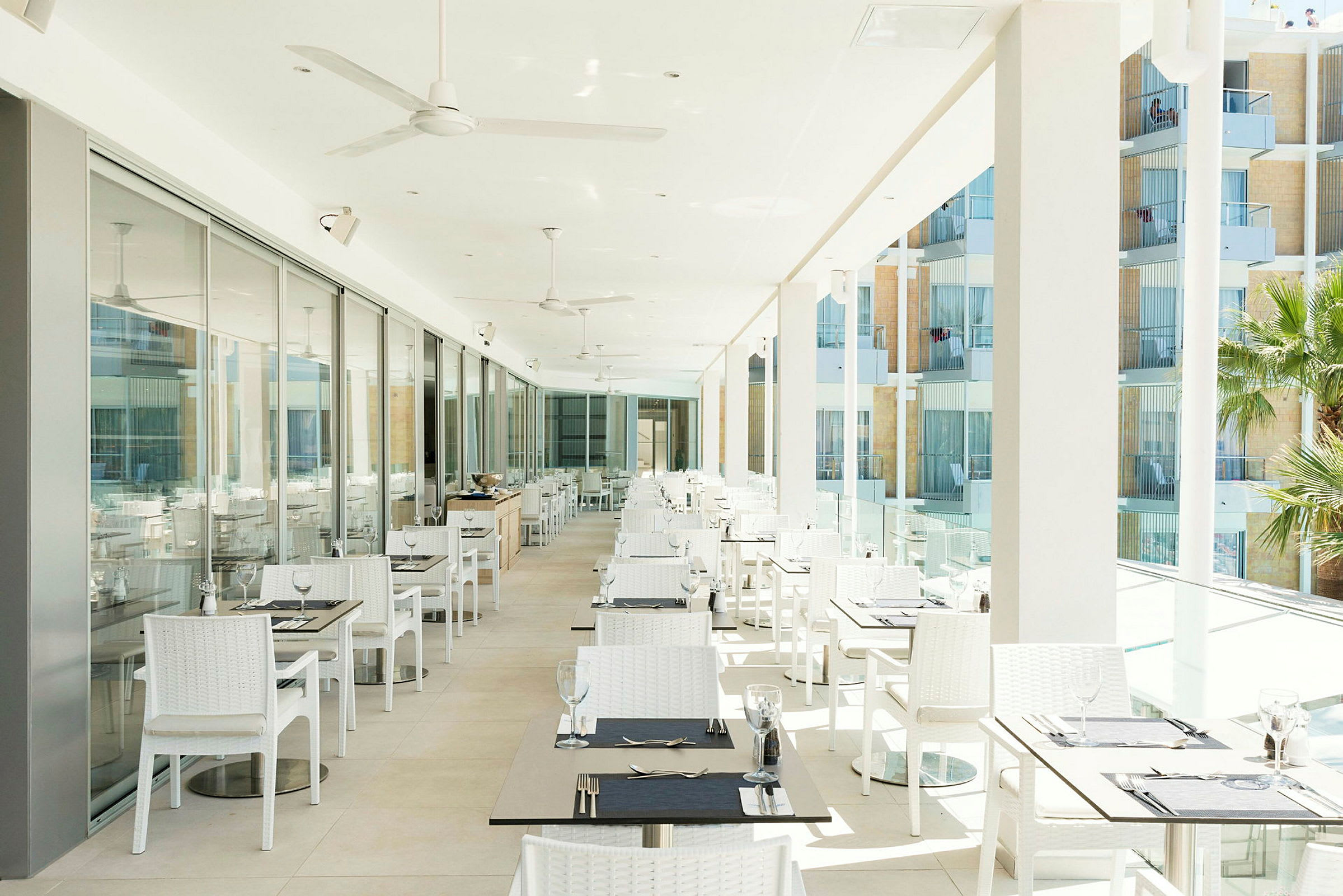 En ljus restaurangterrass med många vita bord och stolar, glasdörrar längs ena sidan och utsikt över balkonger utomhus. (AI-genererad text)