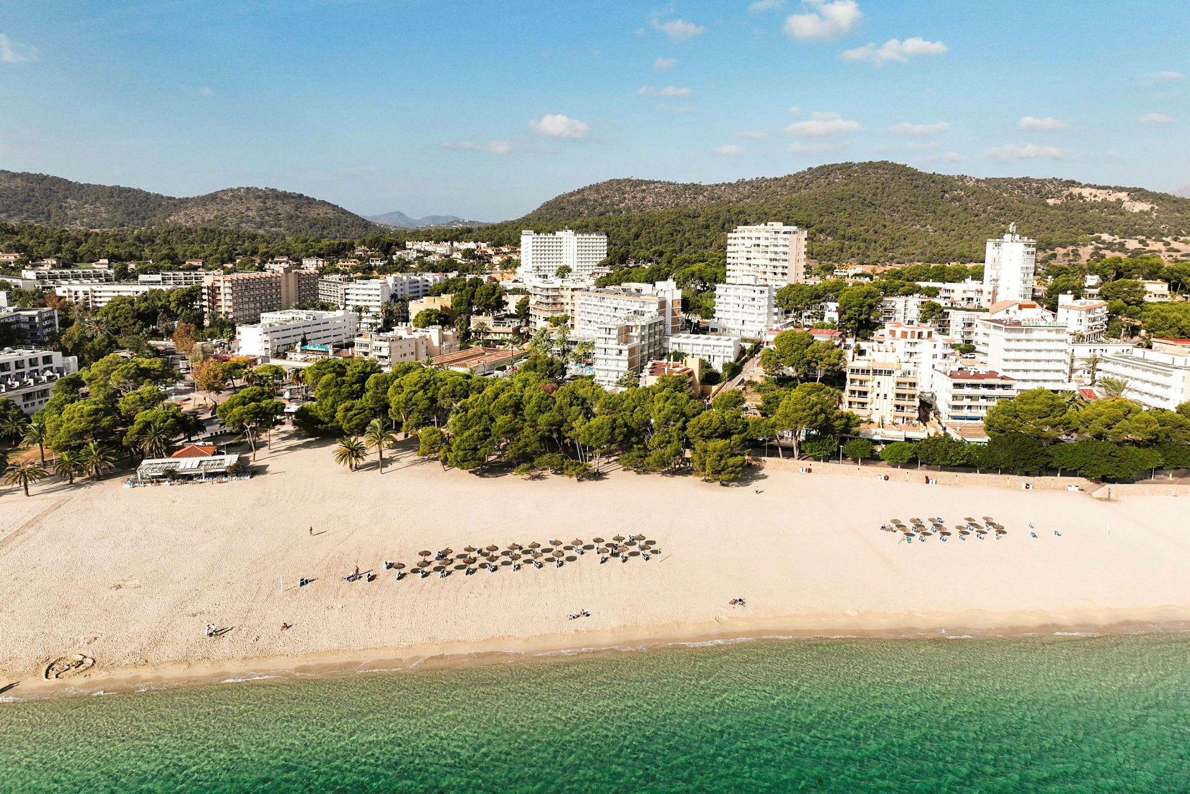 Strand med parasoller och solstolar vid klart vatten, bakom en rad träd och byggnader. (AI-genererad text)