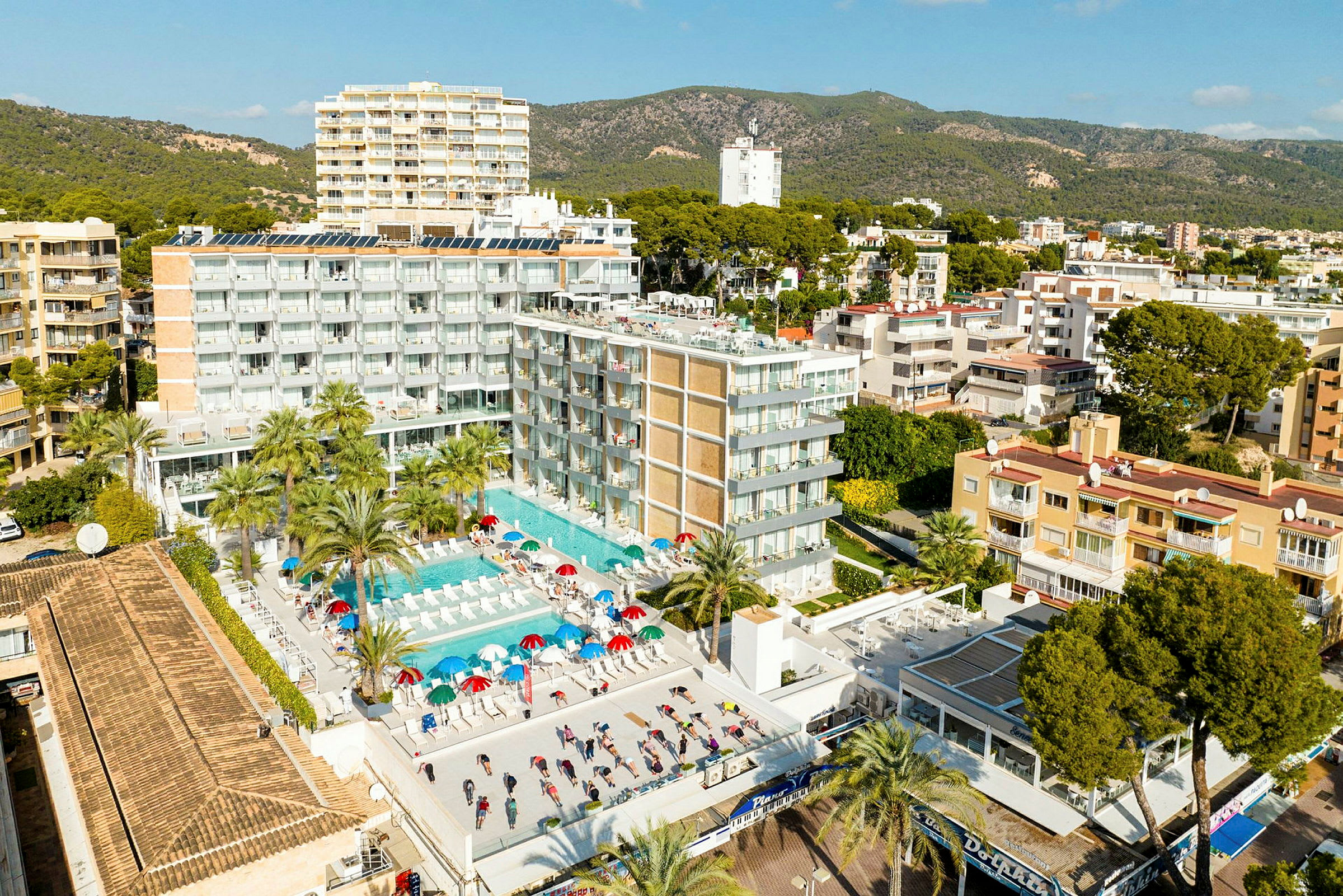 Flygfoto över en hotellbyggnad med poolområde och solstolar. Omgivande landskap med grönska och fler byggnader syns i bakgrunden. (AI-genererad text)
