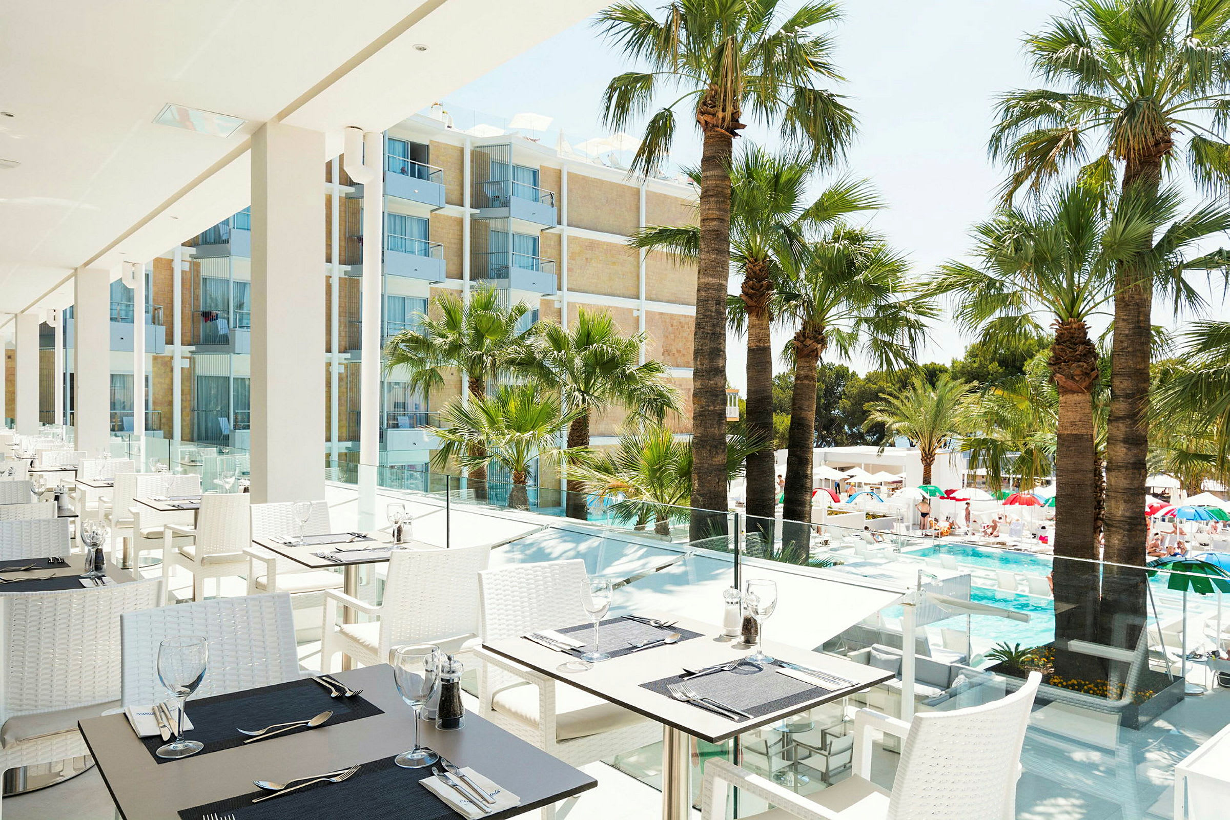 En terrass med uteservering, bord och stolar är placerade nära glasräcken med utsikt över en pool och palmer. I bakgrunden syns en byggnad med balkonger. (AI-genererad text)