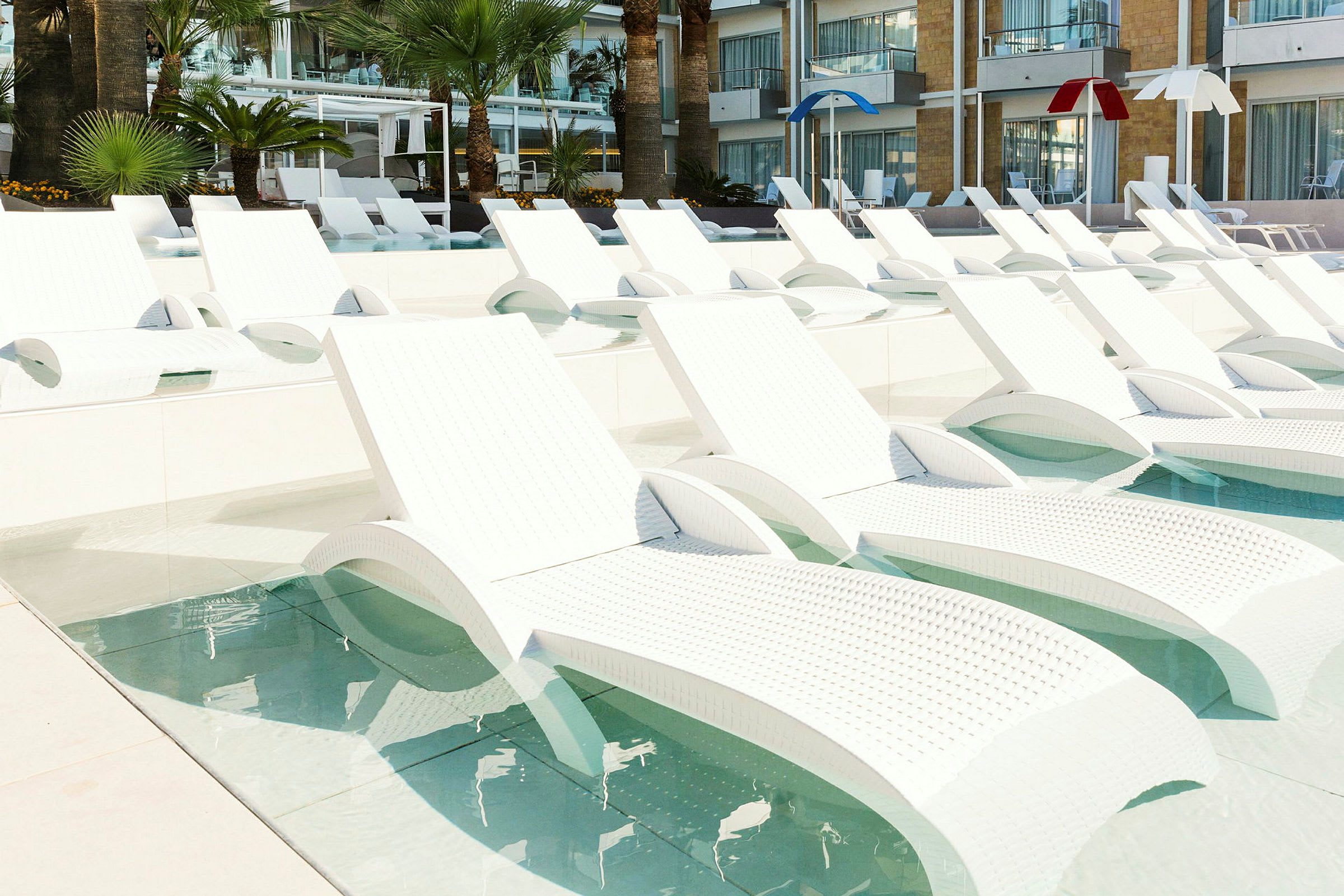 Bild av flera vita solstolar placerade i en grund pool, med palmer och hotellbyggnad i bakgrunden. (AI-genererad text)