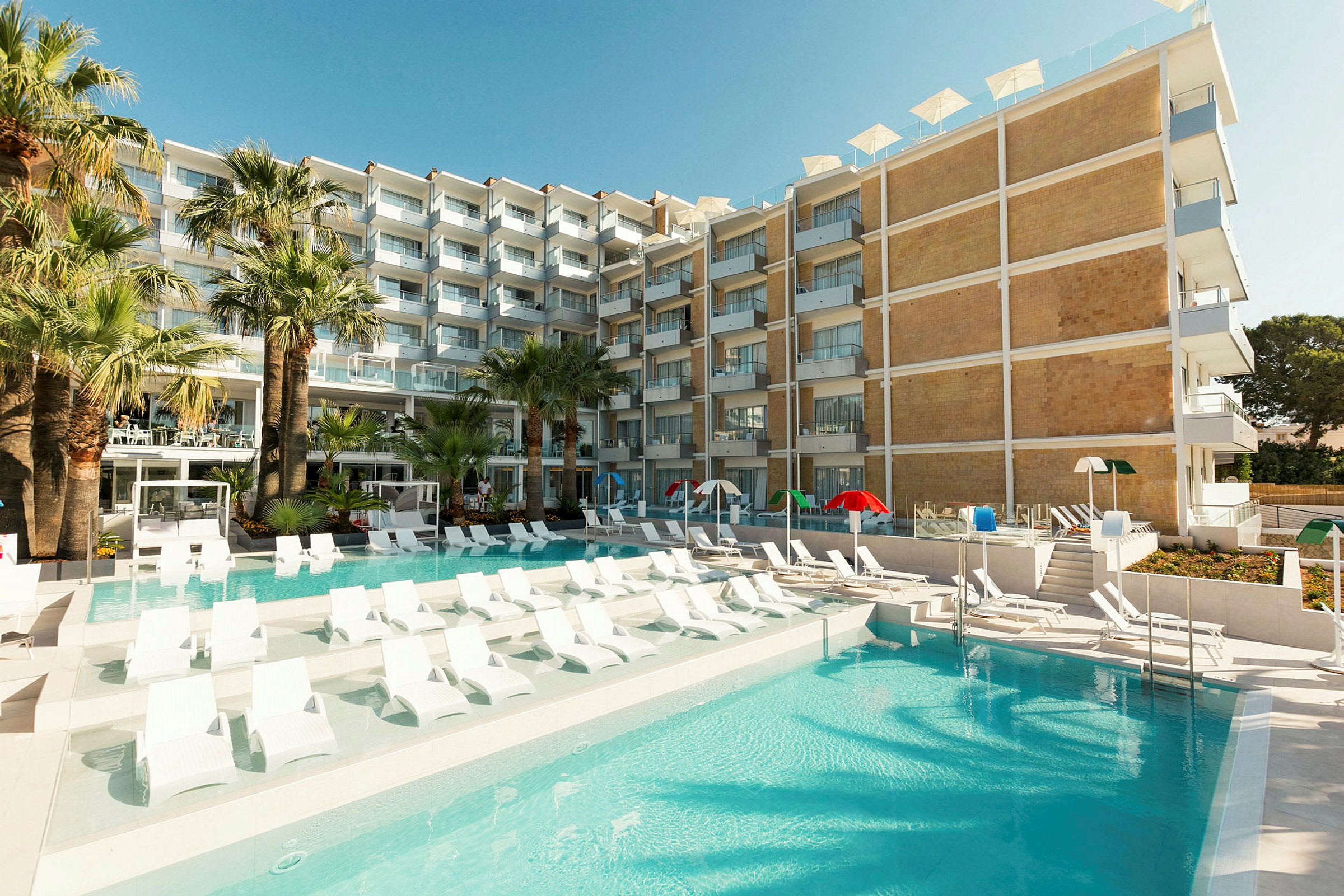 Ett hotell med en pool omgiven av solstolar och gröna palmer. (AI-genererad text)