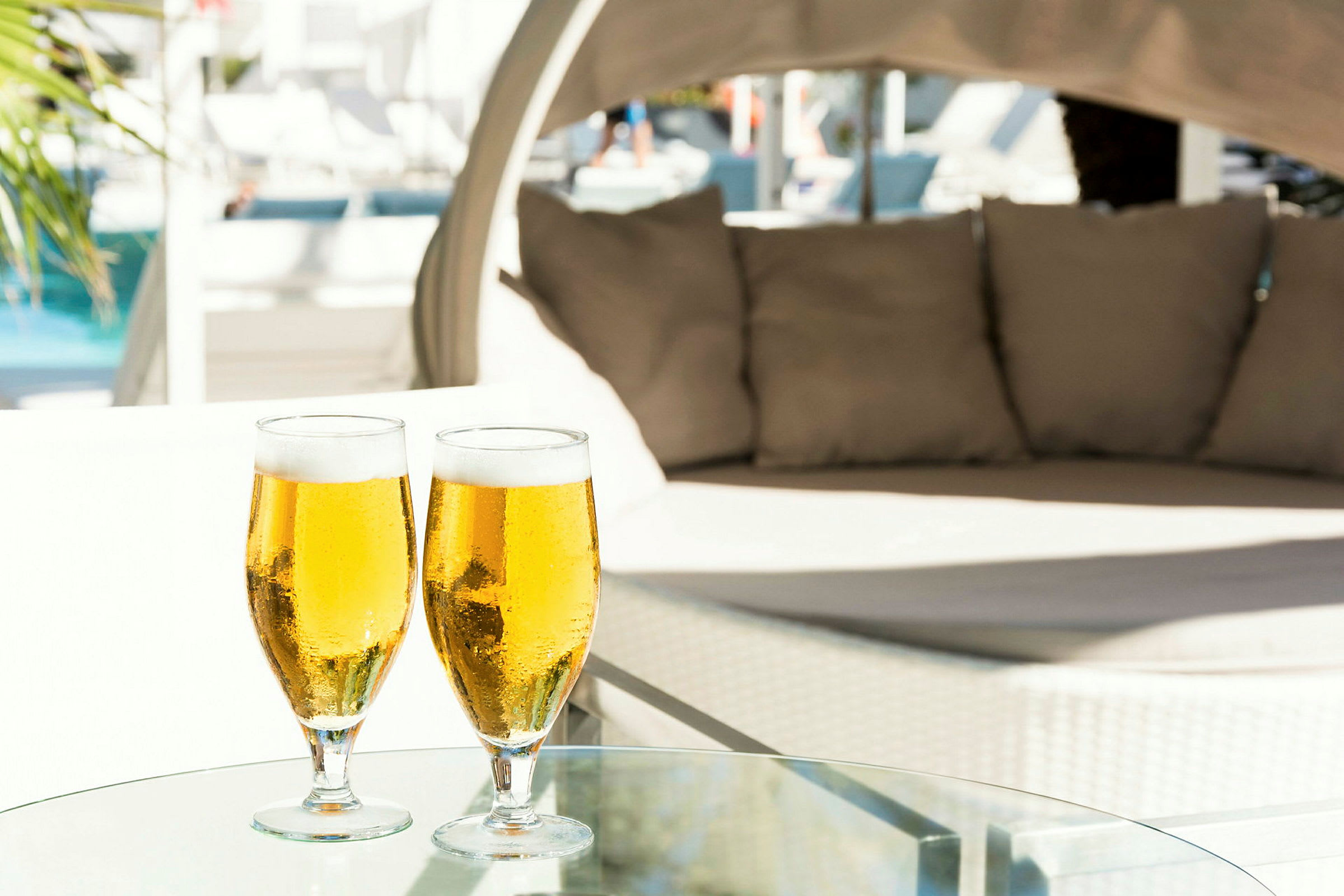 Två glas med öl står på ett glasbord framför en stor utomhusbädd vid en pool. (AI-genererad text)