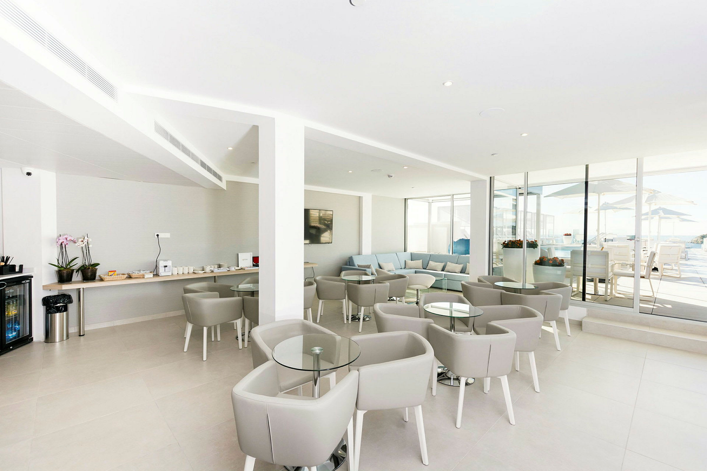 En modern lounge med grå möbler och glasbord. En serveringsyta med drycker och tilltugg är synlig till vänster, och stora glasfönster öppnar upp till en utomhusterrass. (AI-genererad text)