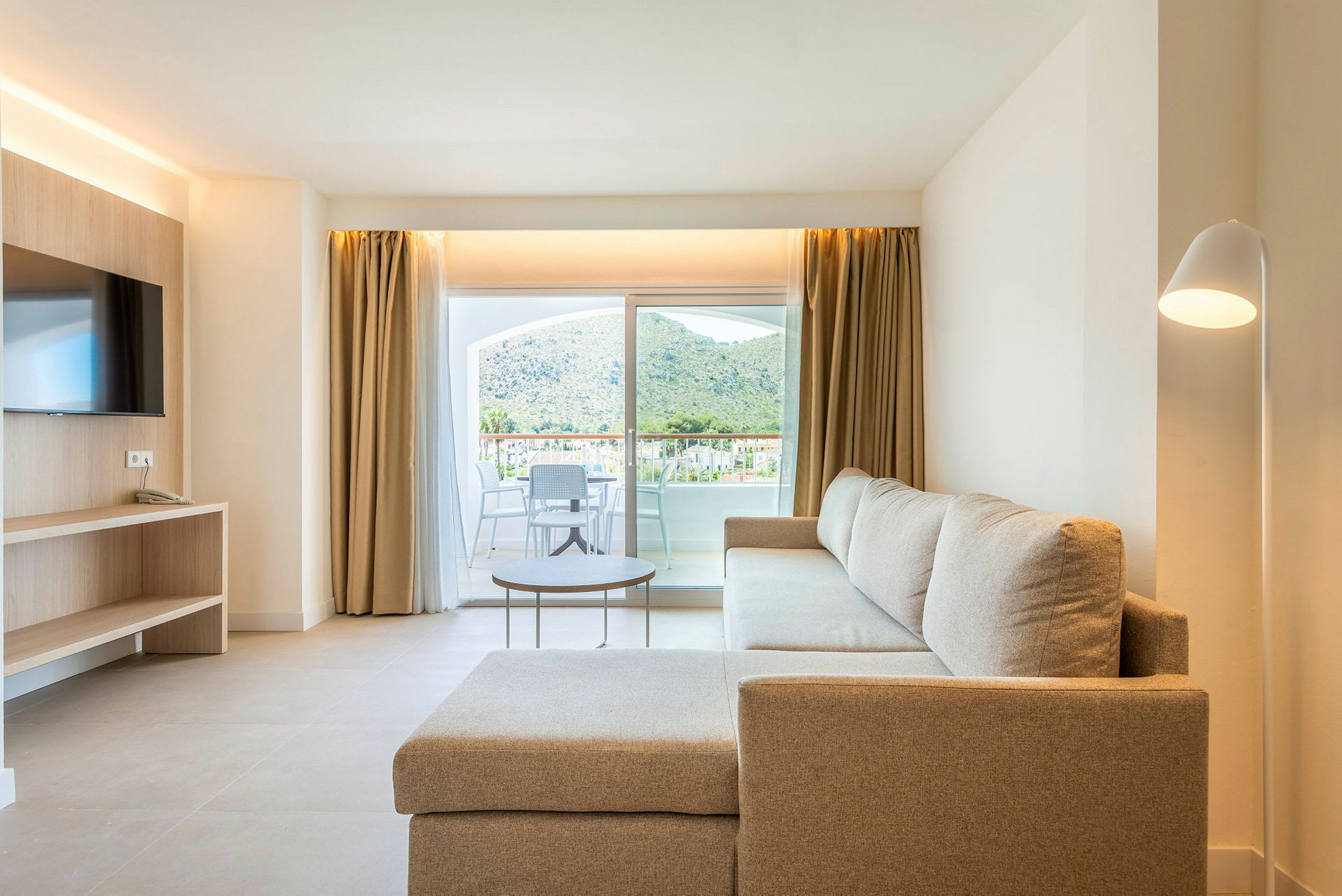 Vardagsrumsinteriör med beige soffa, TV och glasdörr med utsikt mot en terrass och berg. (AI-genererad text)